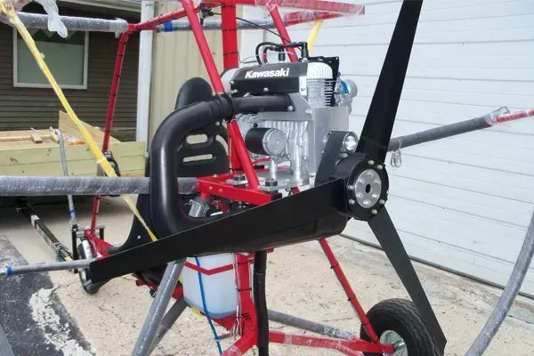 Red framed ultralight aircraft with a Kawasaki engine and black propeller; parked on a paved surface.