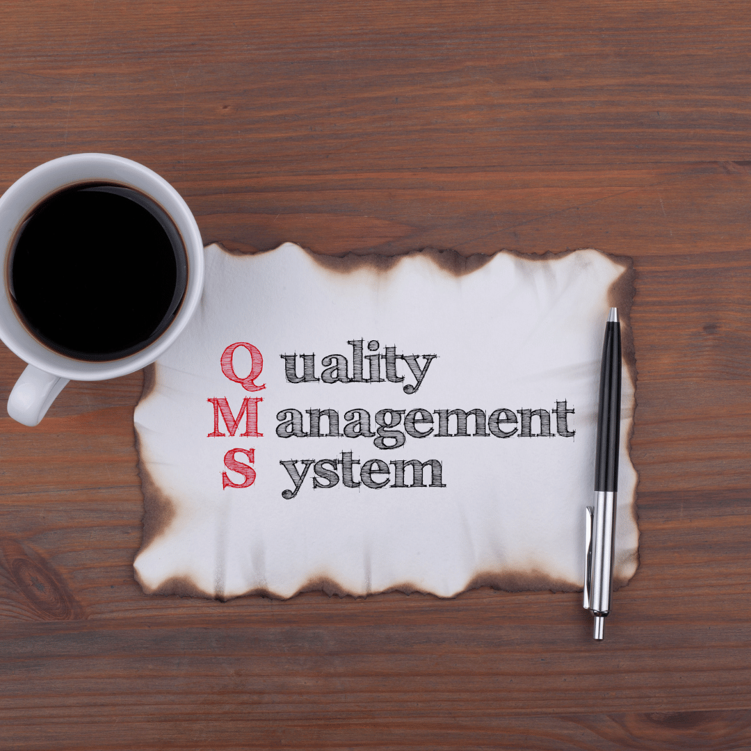 Coffee beside paper reading “Quality management system” on a wooden desk with pens.