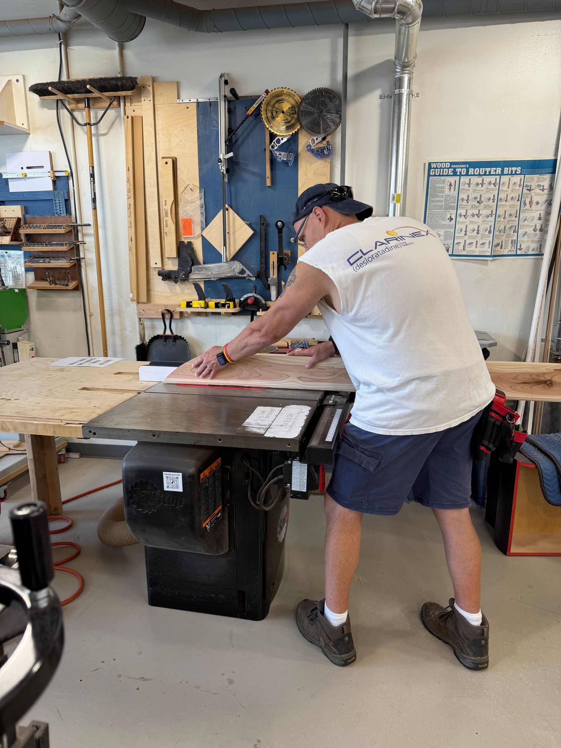 Man using a table saw in a workshop, cutting wood near tools and storage shelves.