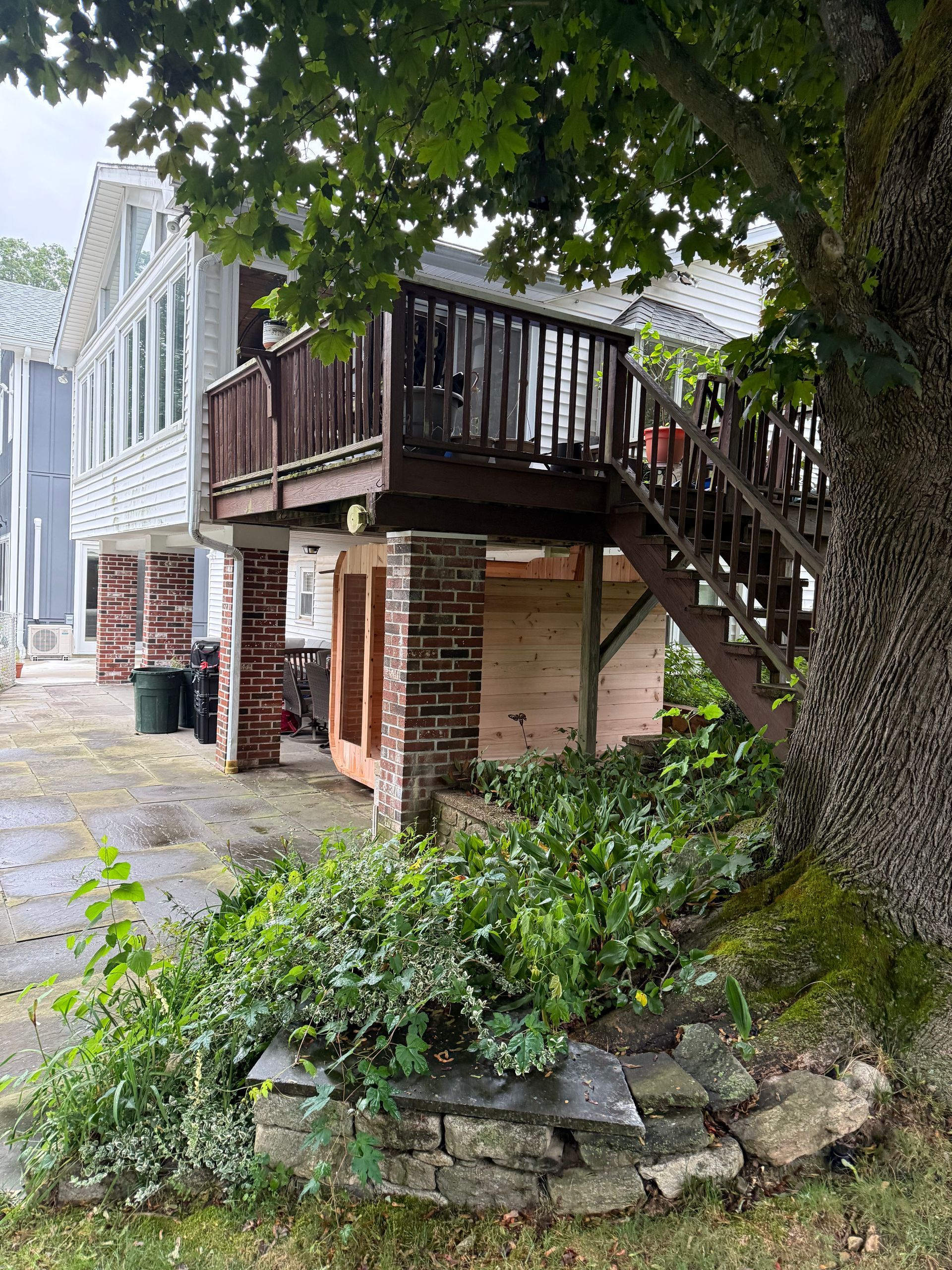 Stone patio under a wooden deck beside a house, with stairs, windows, and greenery in front