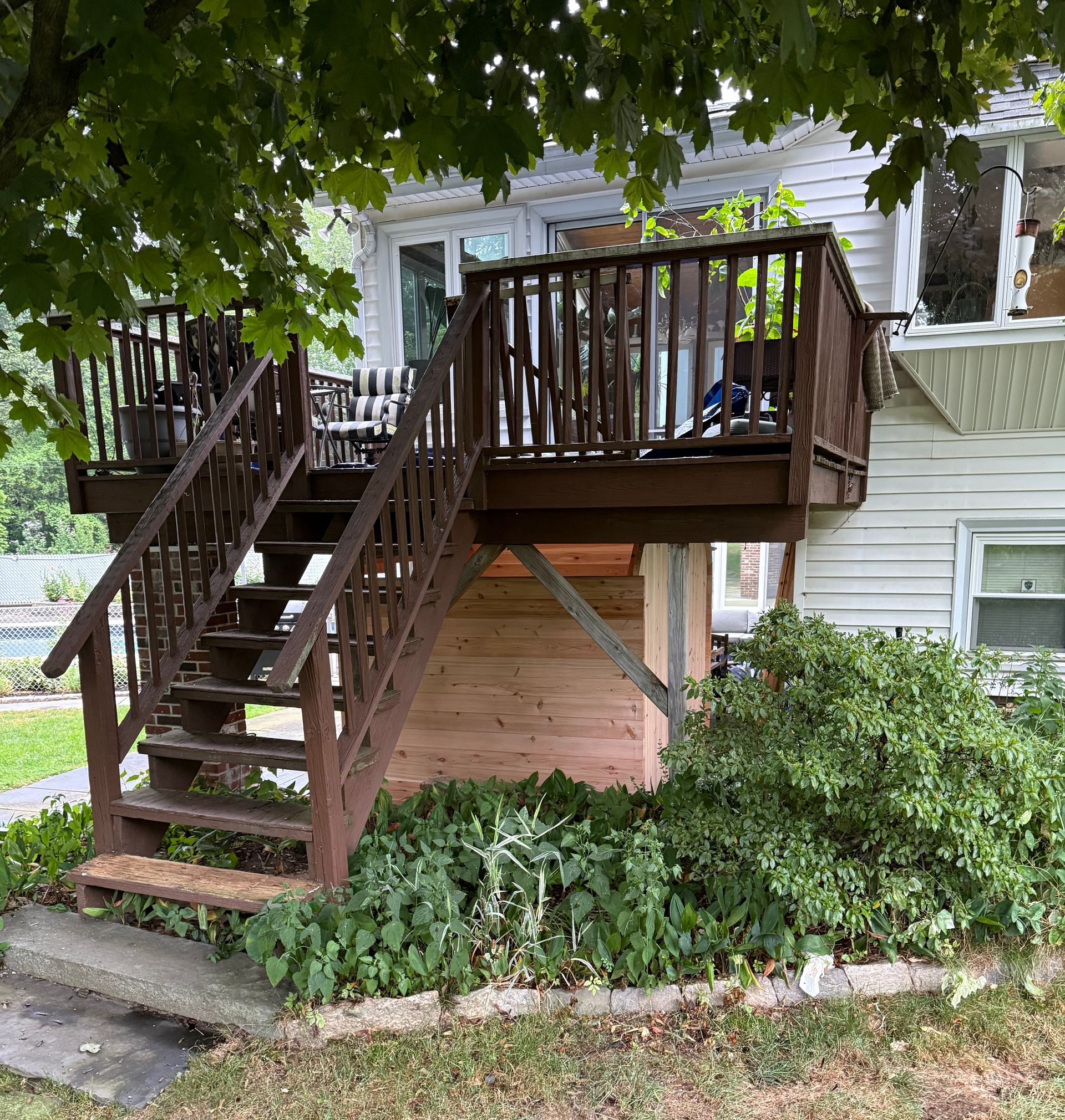 Wooden exterior staircase and deck attached to a house, partly shaded by tree branches and surrounded by greenery.
