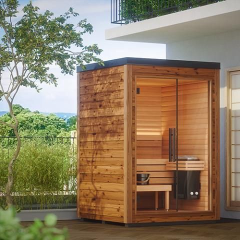 Small wooden outdoor sauna with glass door beside a modern house, surrounded by greenery.