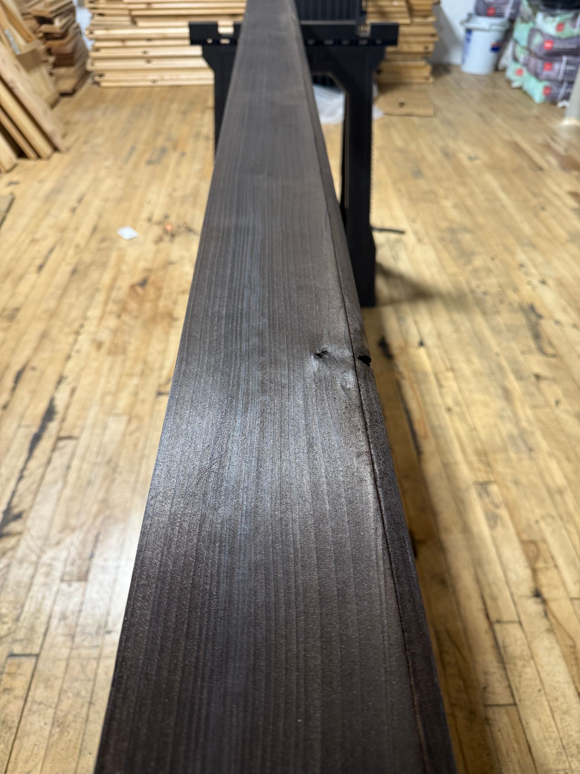 Close-up of a long dark wooden beam on a workshop floor with stacks of lumber in the background