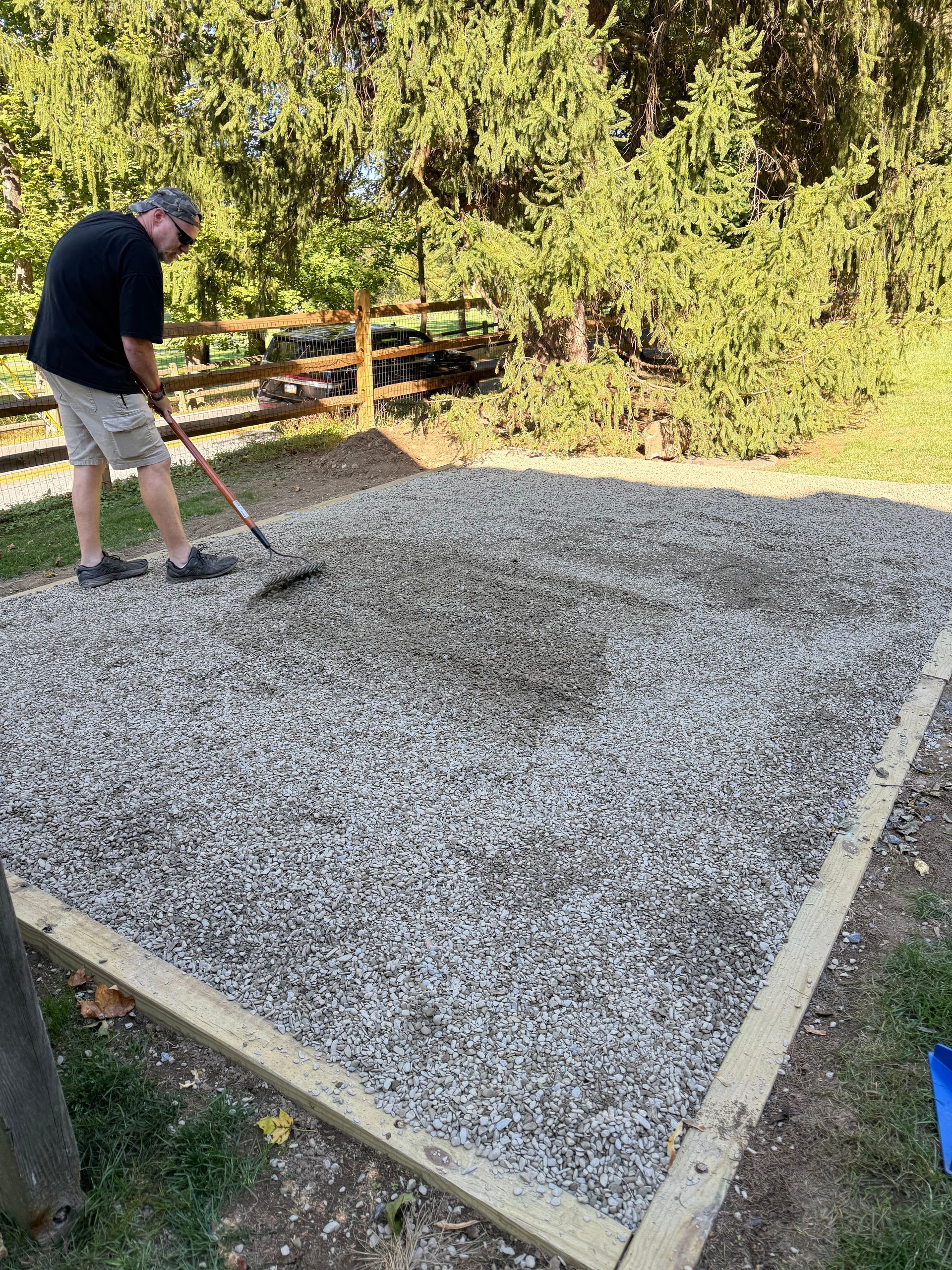 Man spreading gravel over a framed concrete pad outdoors with a shovel
