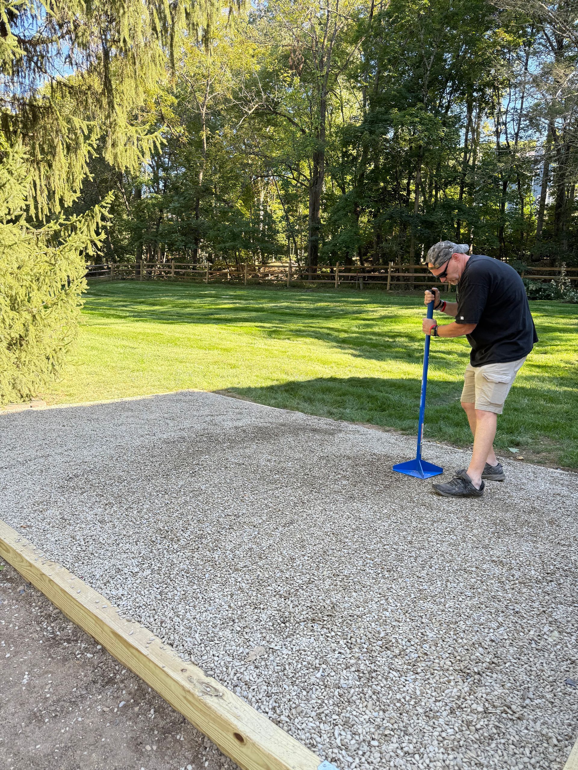 Person using a blue post-hole digger on a gravel area beside a grassy yard.