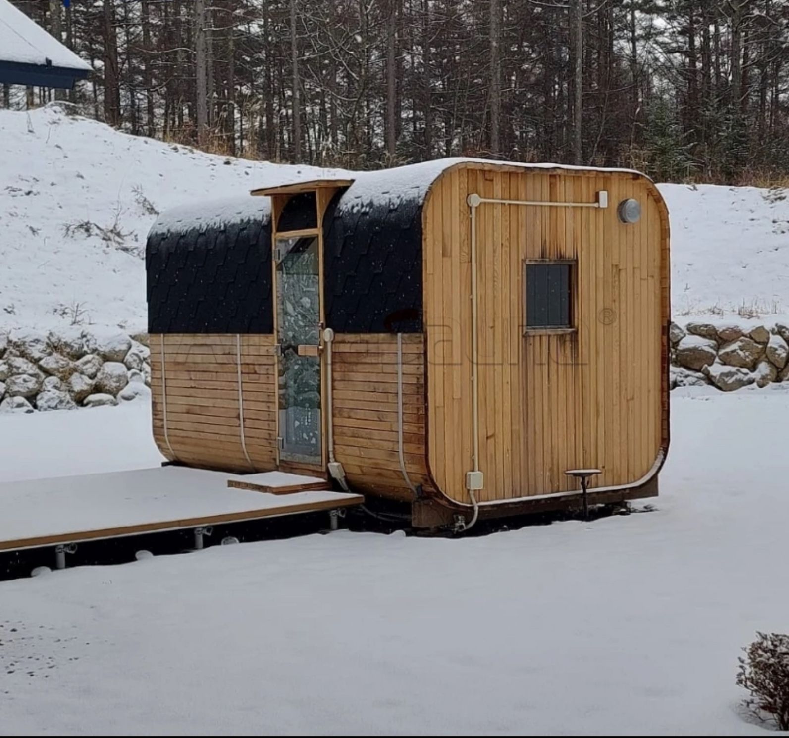 Small wooden cabin with a black curved roof sitting on snow beside a forest.