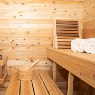 Wood-paneled sauna with wooden benches, bucket and ladle, and rolled towels on a bench.