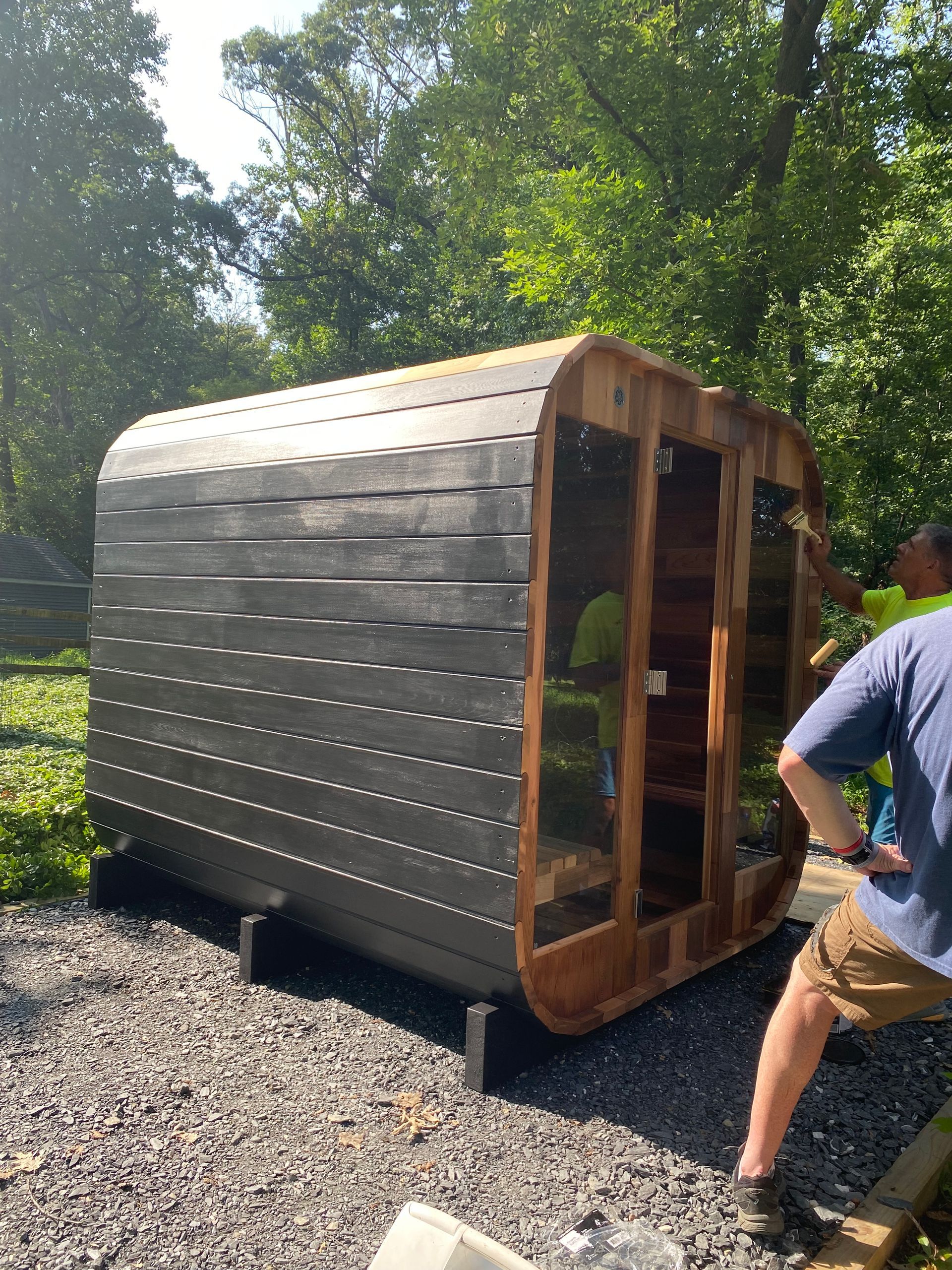 Wooden garden sauna or shed on gravel, with a person standing beside it in a leafy yard.