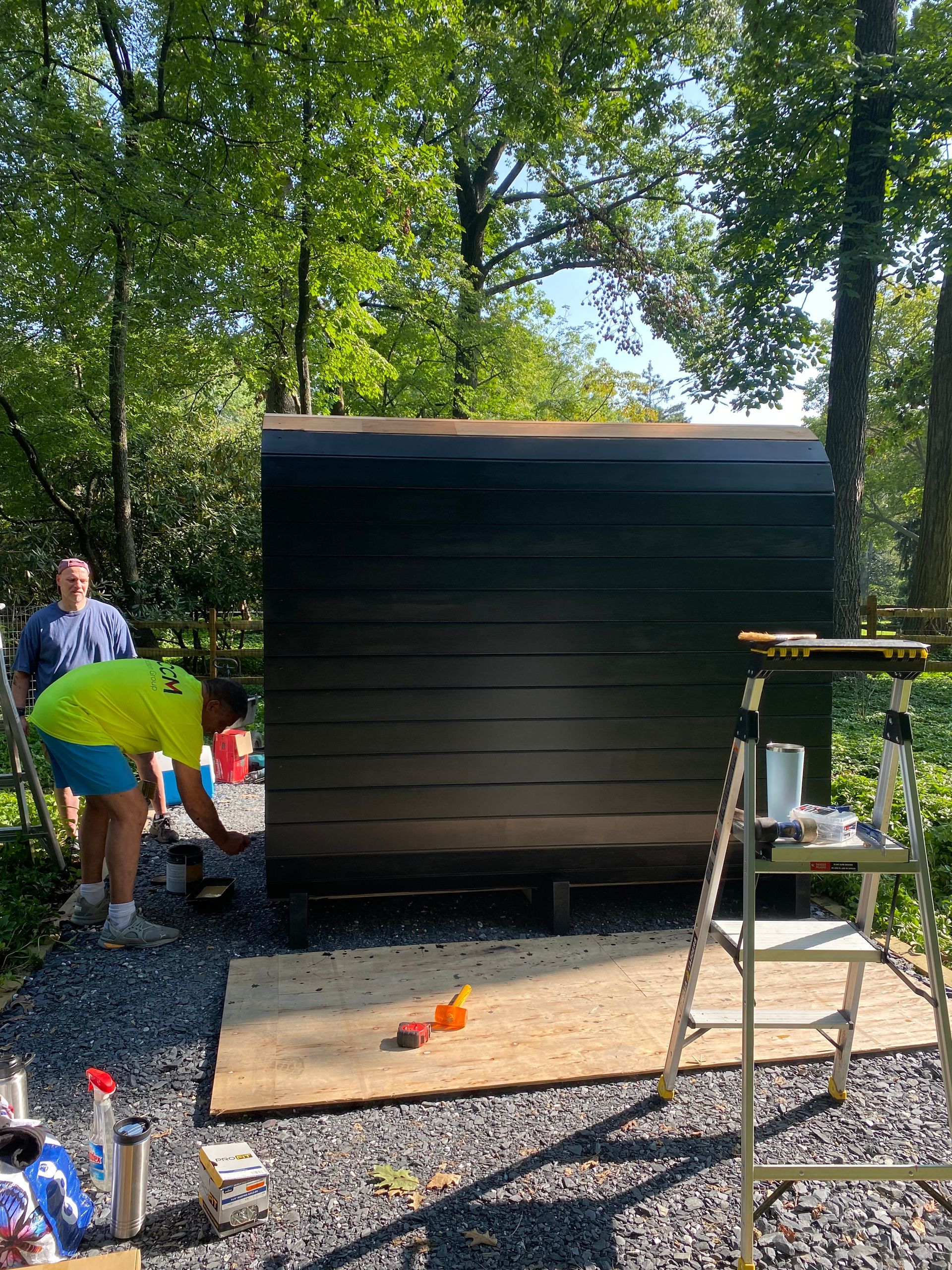 Two people assembling a dark wood shed outdoors among trees and tools.