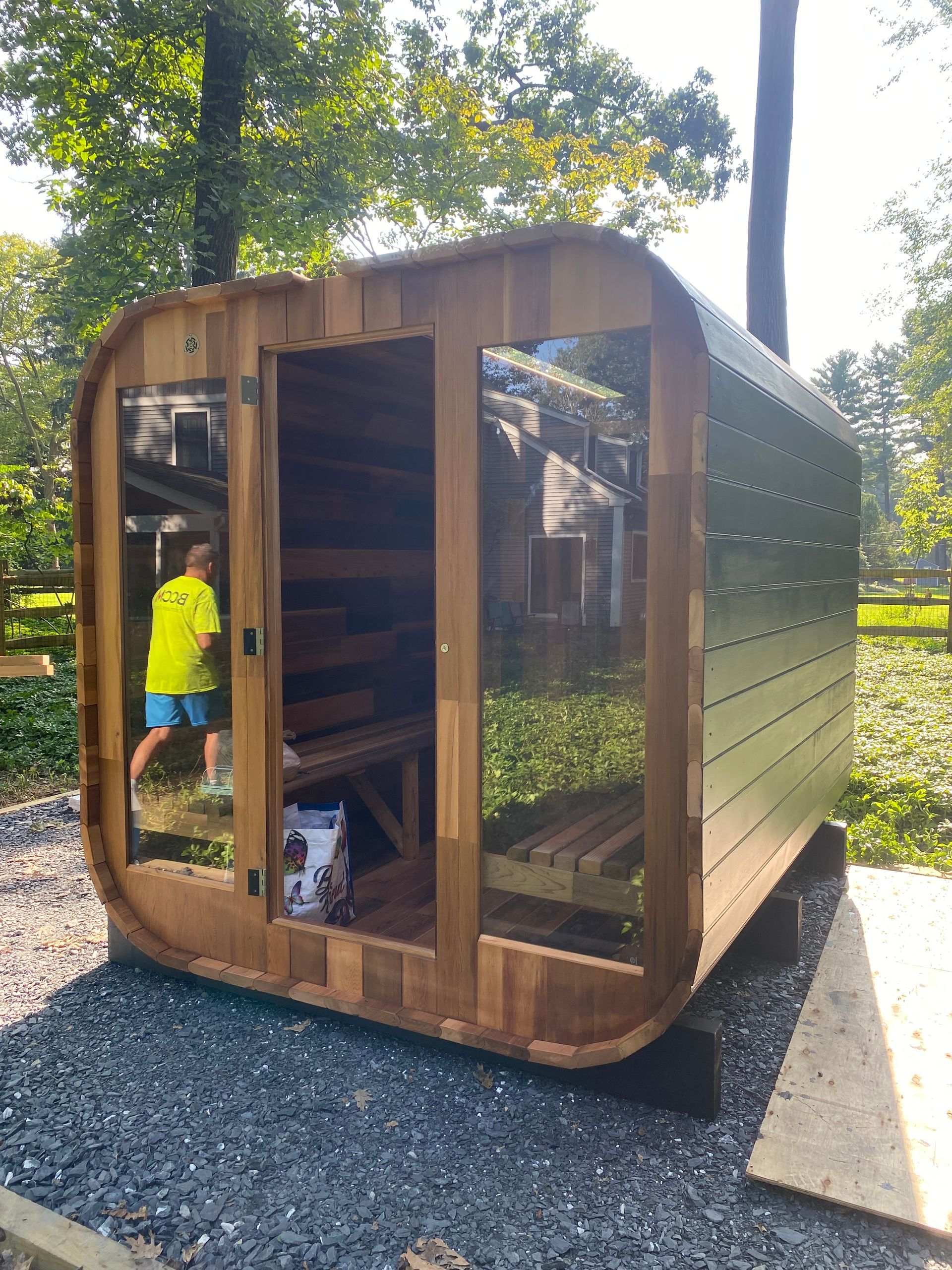 Small wooden garden shed with rounded front, a person in a yellow shirt inside, surrounded by trees and gravel.