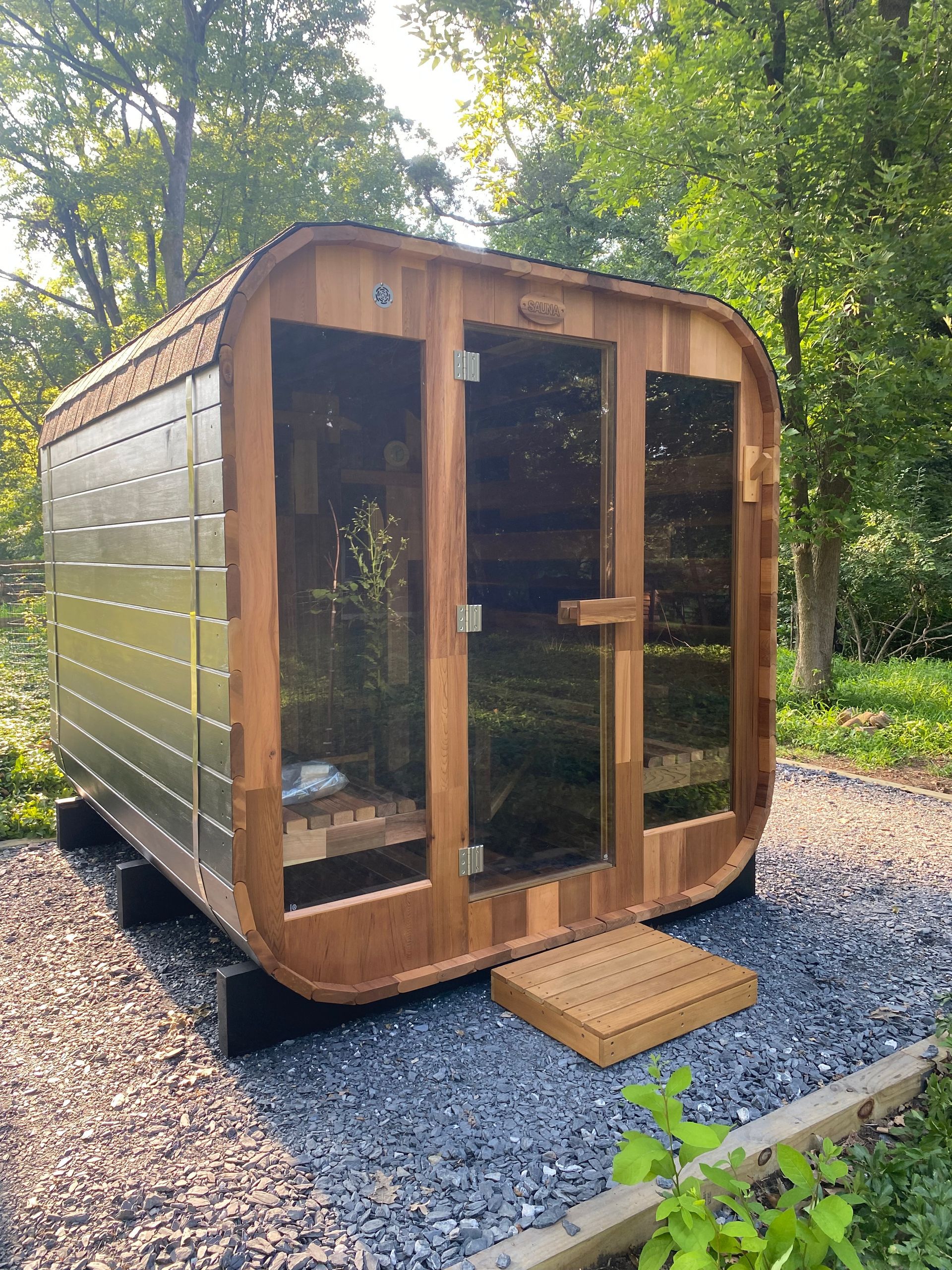 Small wooden sauna cabin with glass doors on a gravel pad in a wooded backyard
