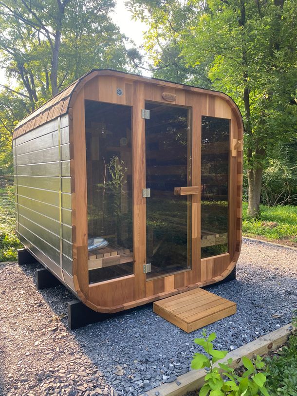 Small wooden sauna cabin with glass door and windows, set on gravel in a wooded outdoor area