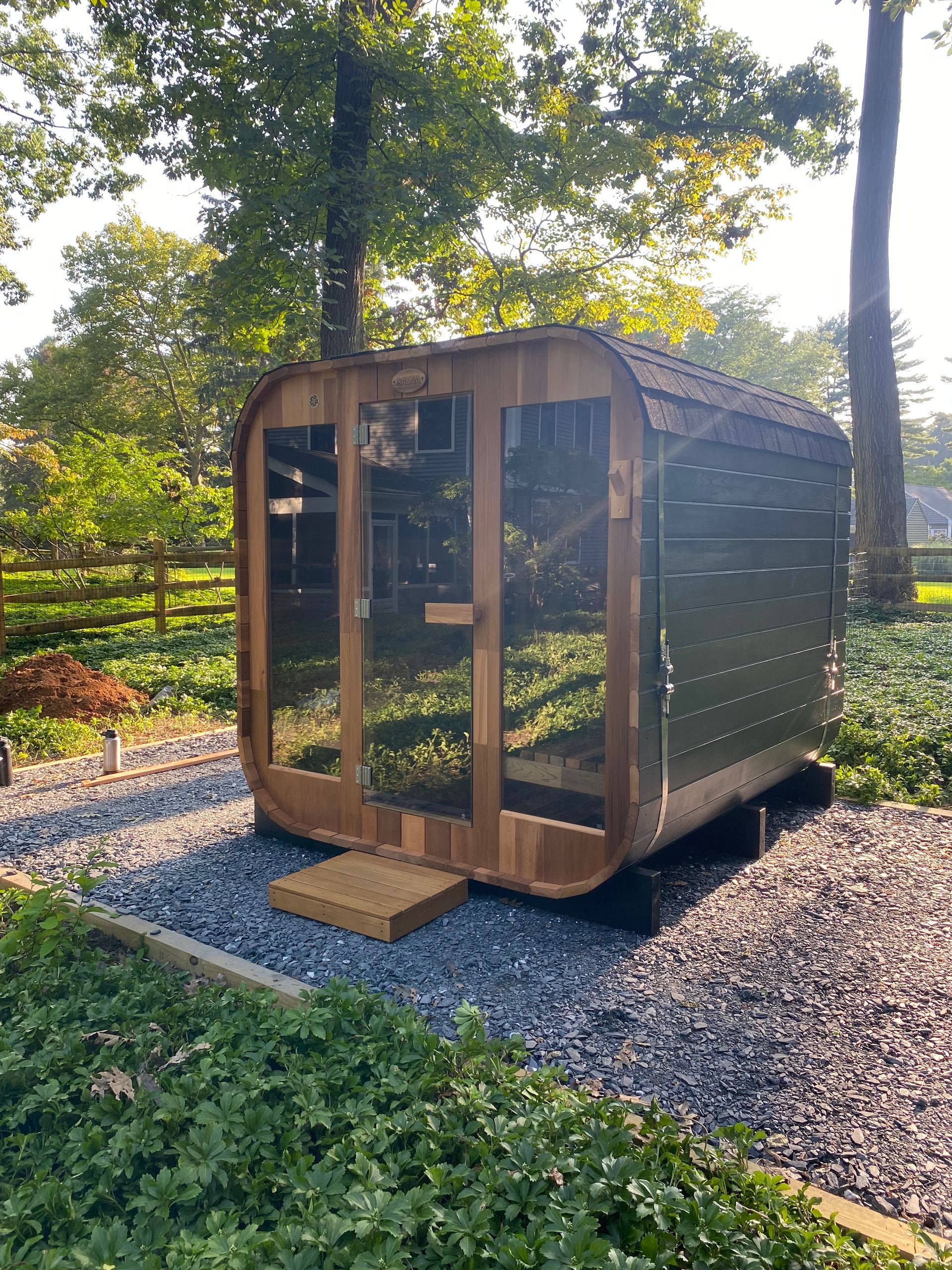 Small wooden sauna cabin with glass doors on gravel in a wooded yard