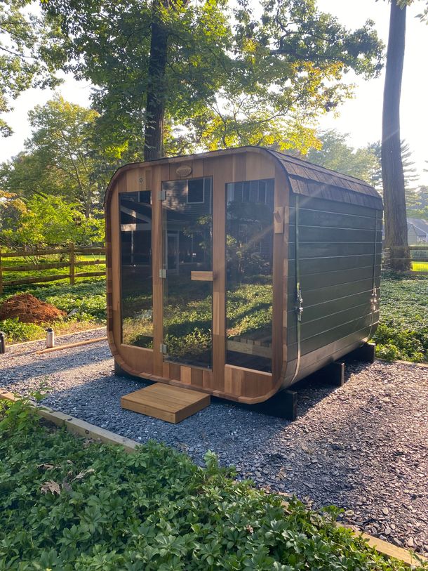 Small wooden sauna cabin with glass front, set on gravel in a wooded yard with sunlight filtering through trees