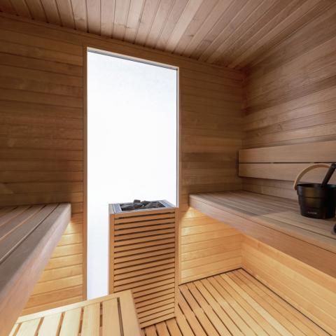 Wood-paneled sauna interior with benches, slatted floor, and a bucket near a bright window.