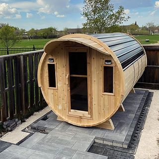 Wooden 4 person barrel-shaped outdoor sauna with glass door on a paved patio in a yard