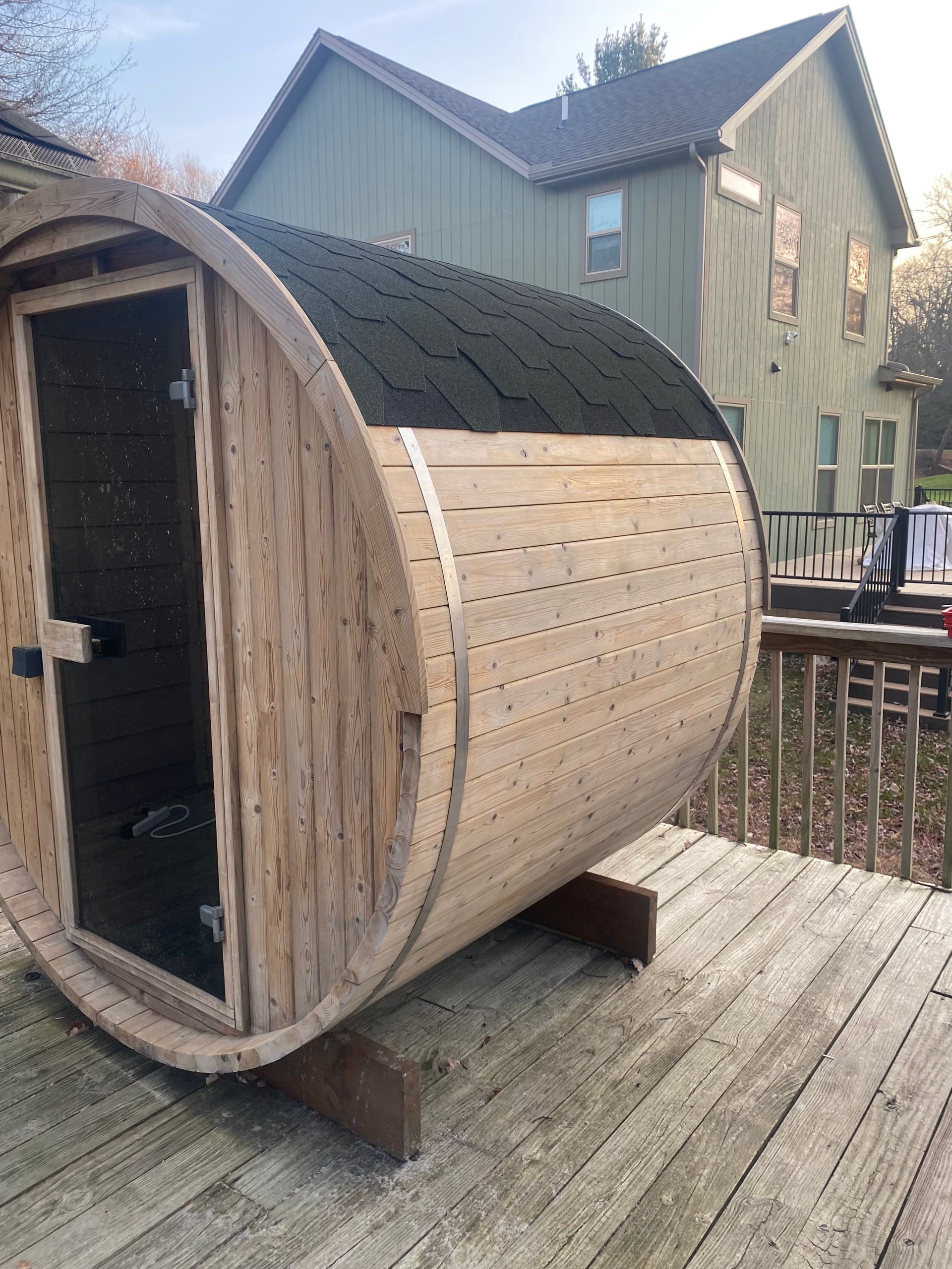 Wooden barrel-shaped sauna pod on a deck beside a house, with a small open doorway.
