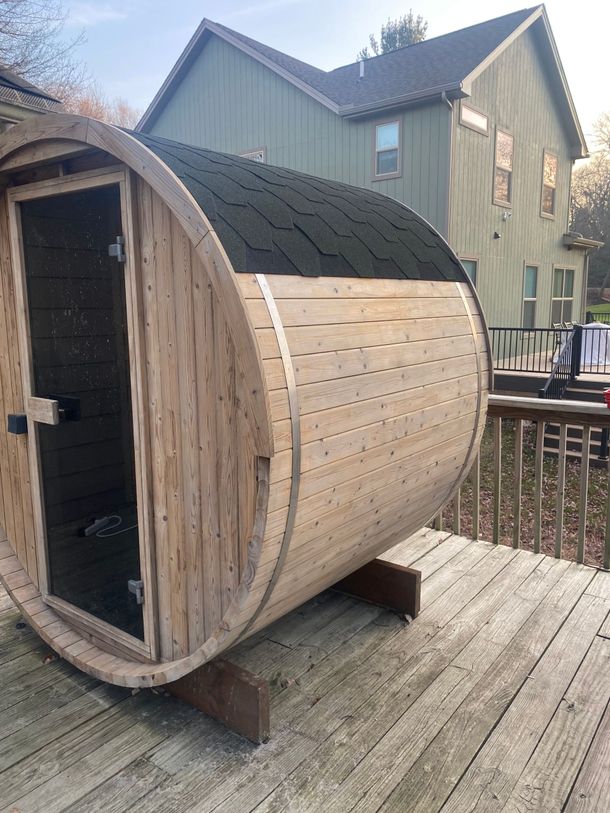 Wooden barrel-shaped sauna pod on a deck beside a house, with a small open doorway.