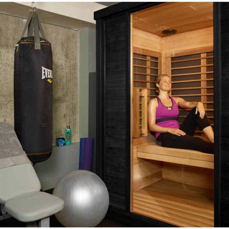 Woman seated in a wooden sauna beside a gym area with a punching bag and exercise ball