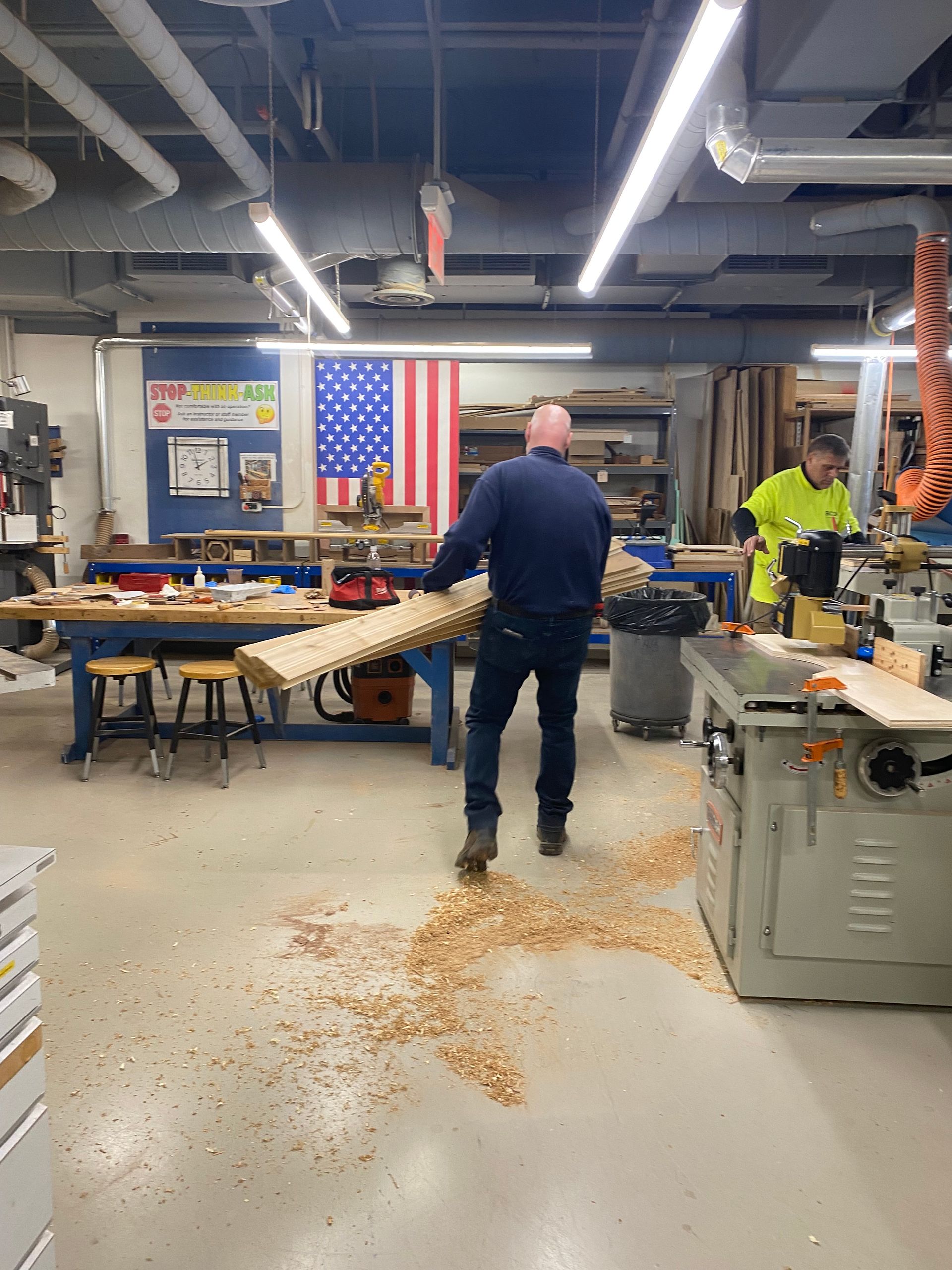 Workshop with tables and tools; a person walks through spilled wood shavings on the floor.