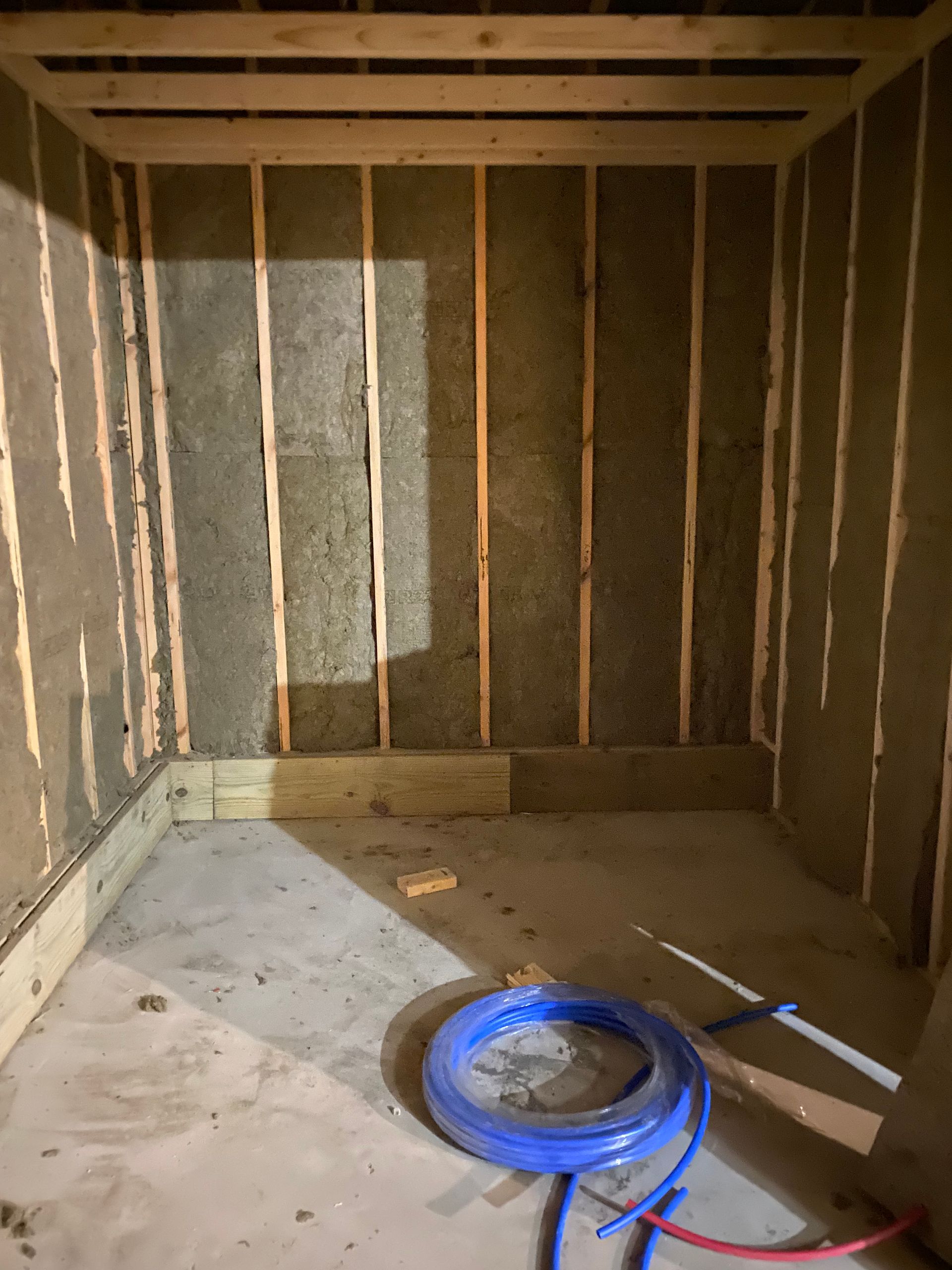 Empty unfinished room with exposed studs, concrete floor, and a blue coiled hose near the center.