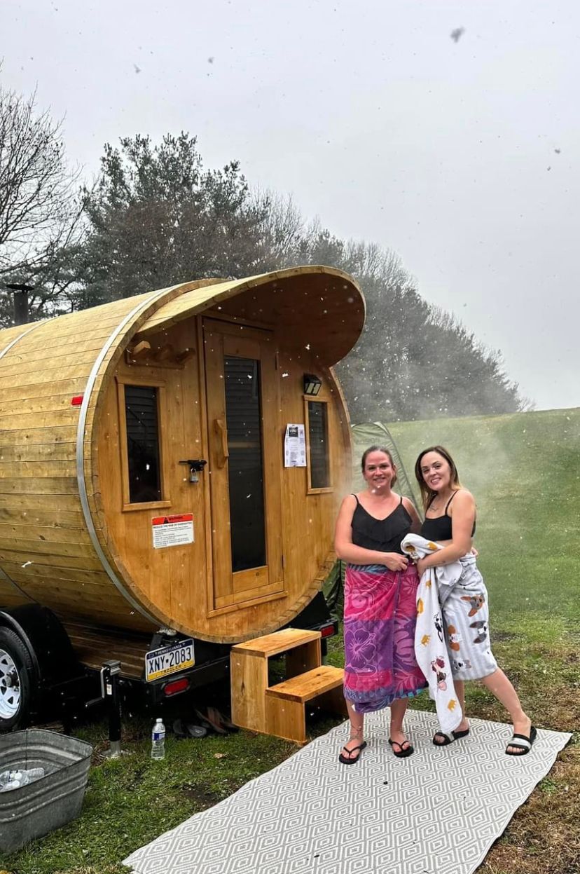 Two people standing beside a small wooden trailer in a grassy field with mist in the background