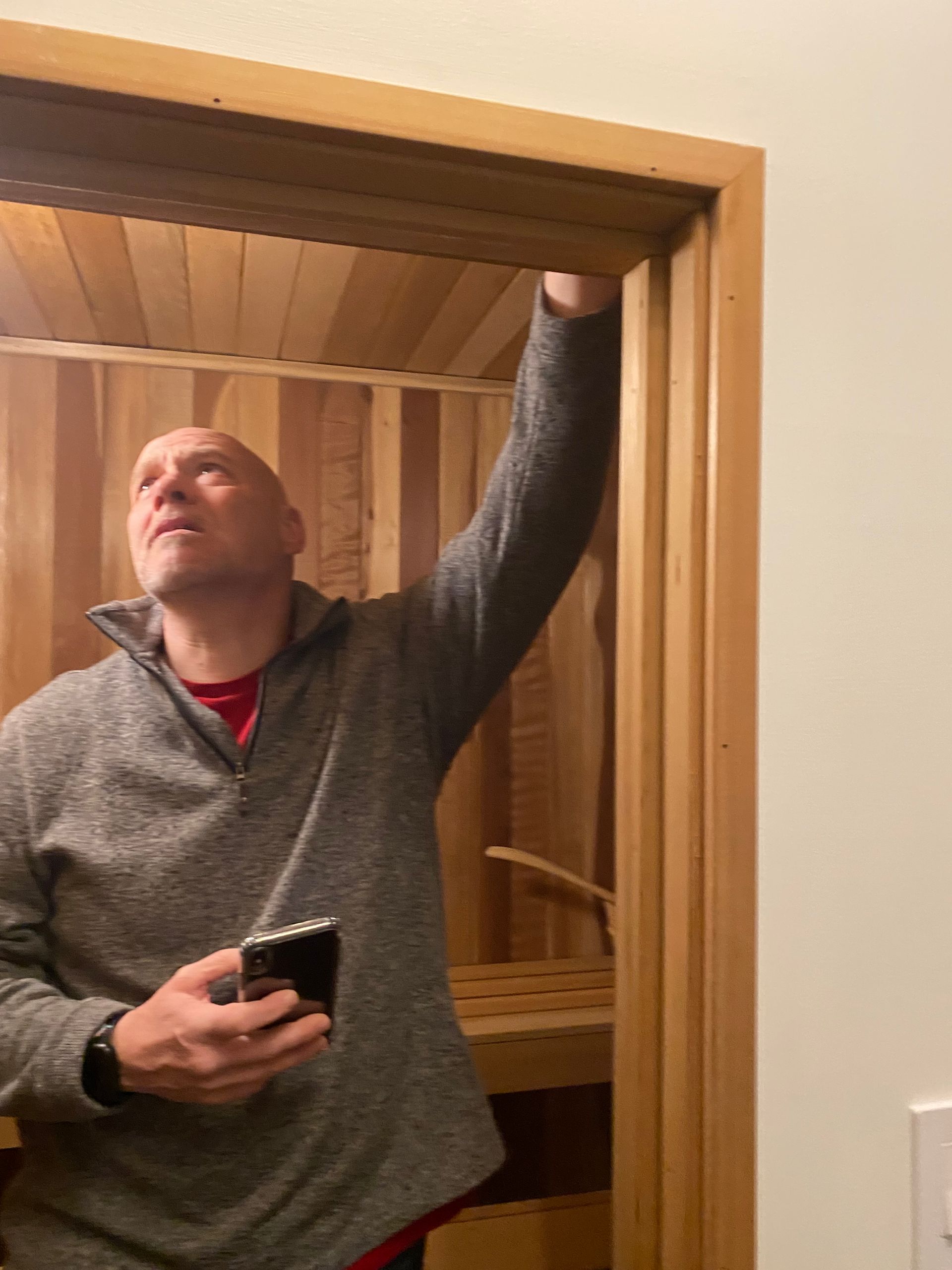 Man in a gray sweater reaching up in a doorway, holding a phone, inside a wooden room