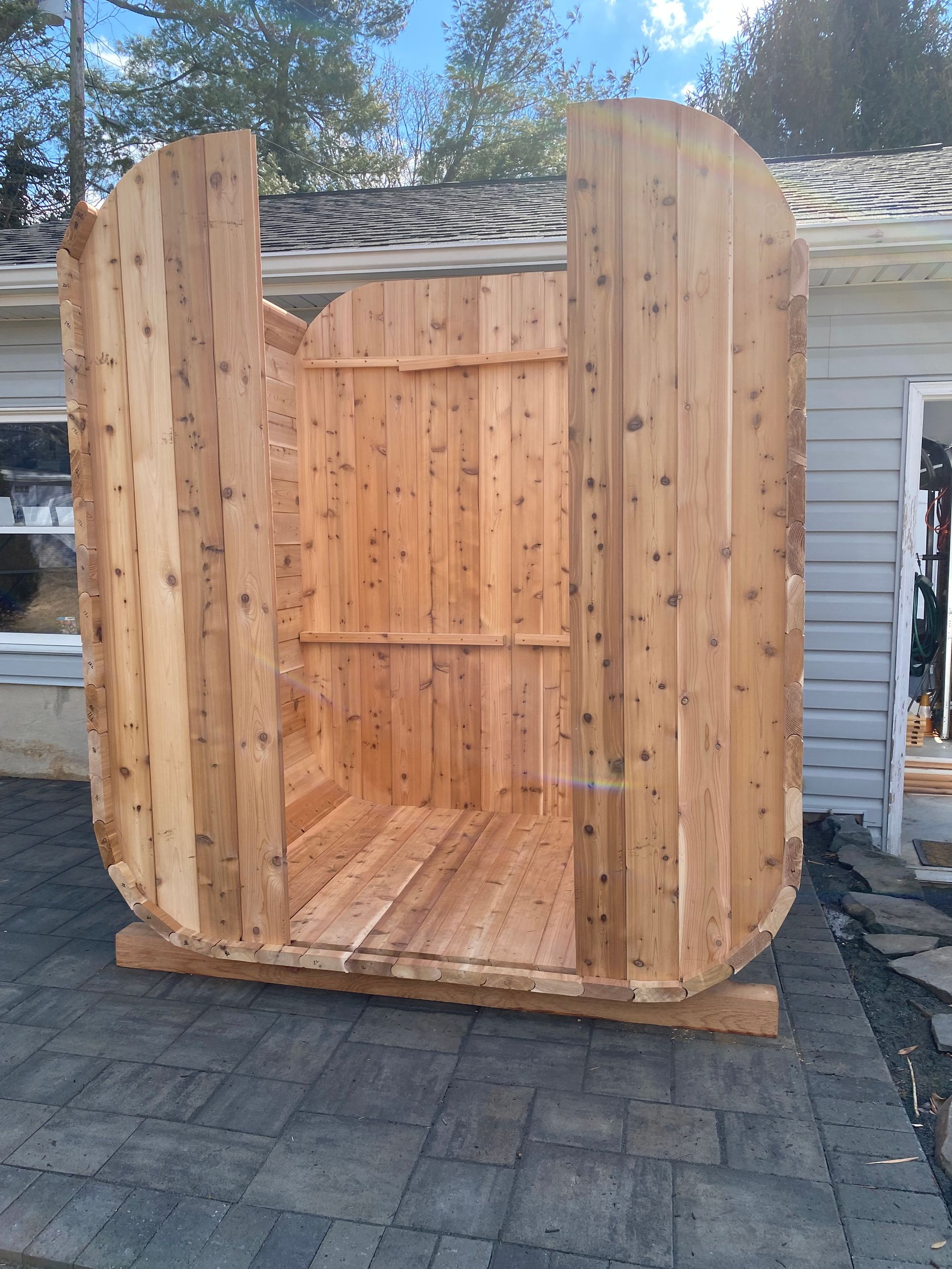 Wooden barrel-shaped sauna pod with open front, standing on a patio beside a house.