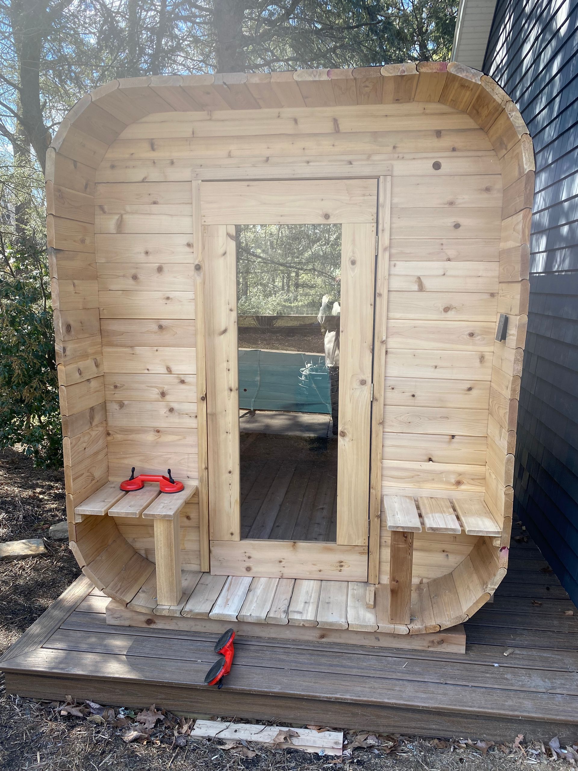 Small unfinished wooden playhouse with a doorway, steps, and benches outdoors among trees.