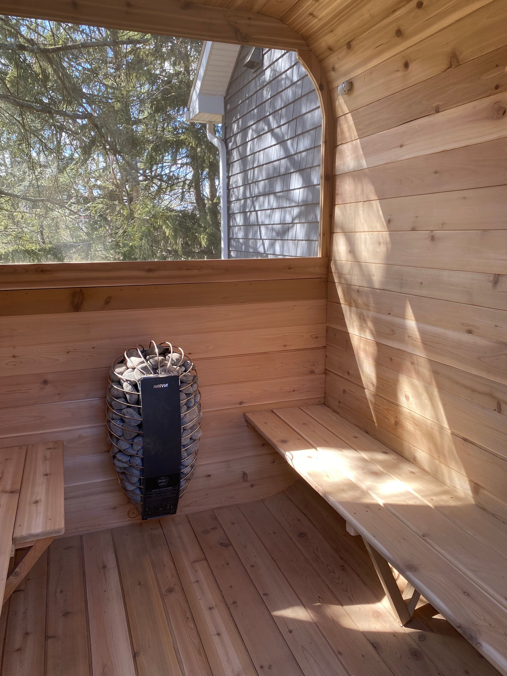 Wooden sauna interior with bench seating and a heater on the floor, sunlight streaming through the window.
