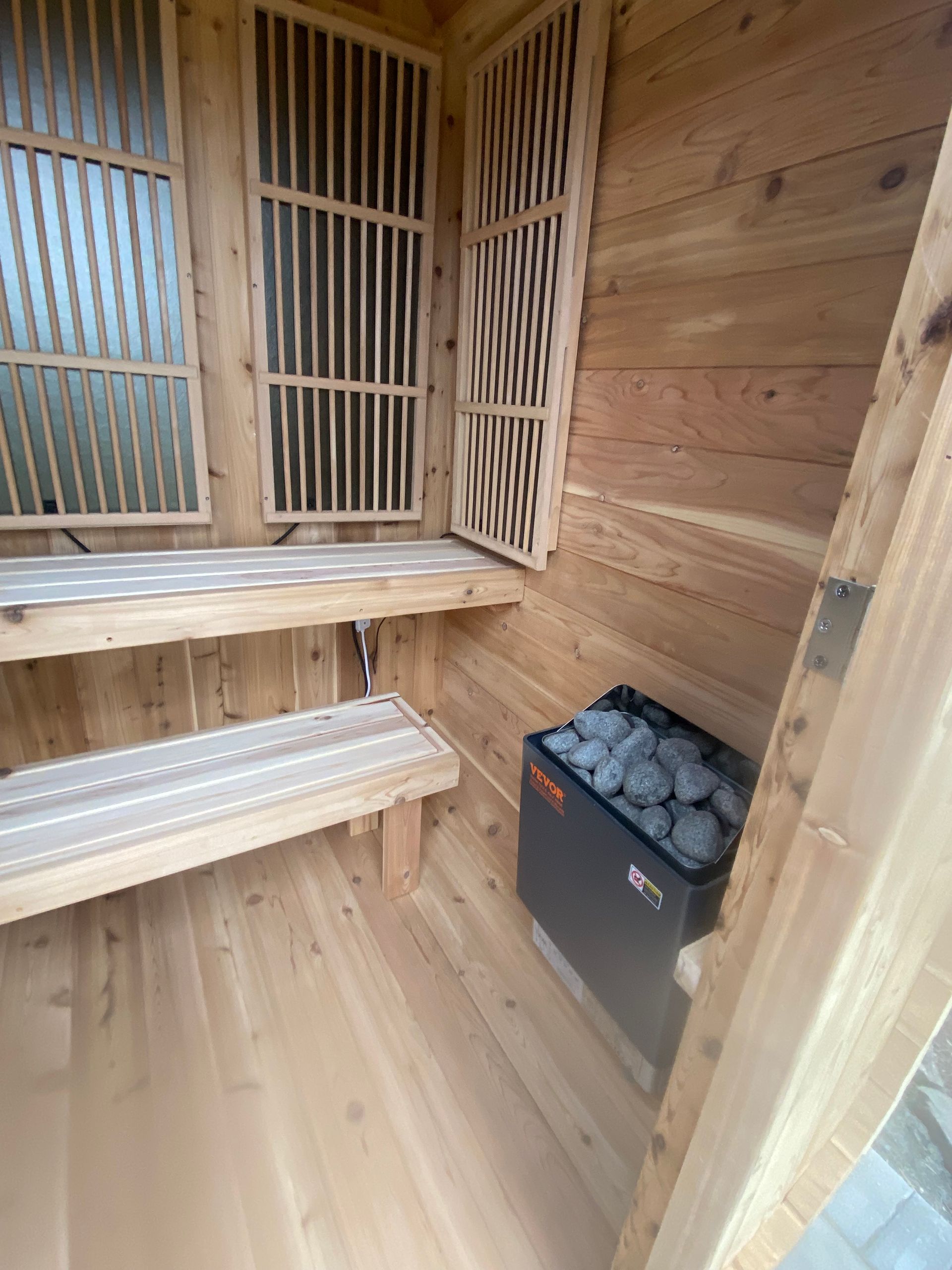 Small wooden sauna cabin with glass doors on gravel in a wooded yard