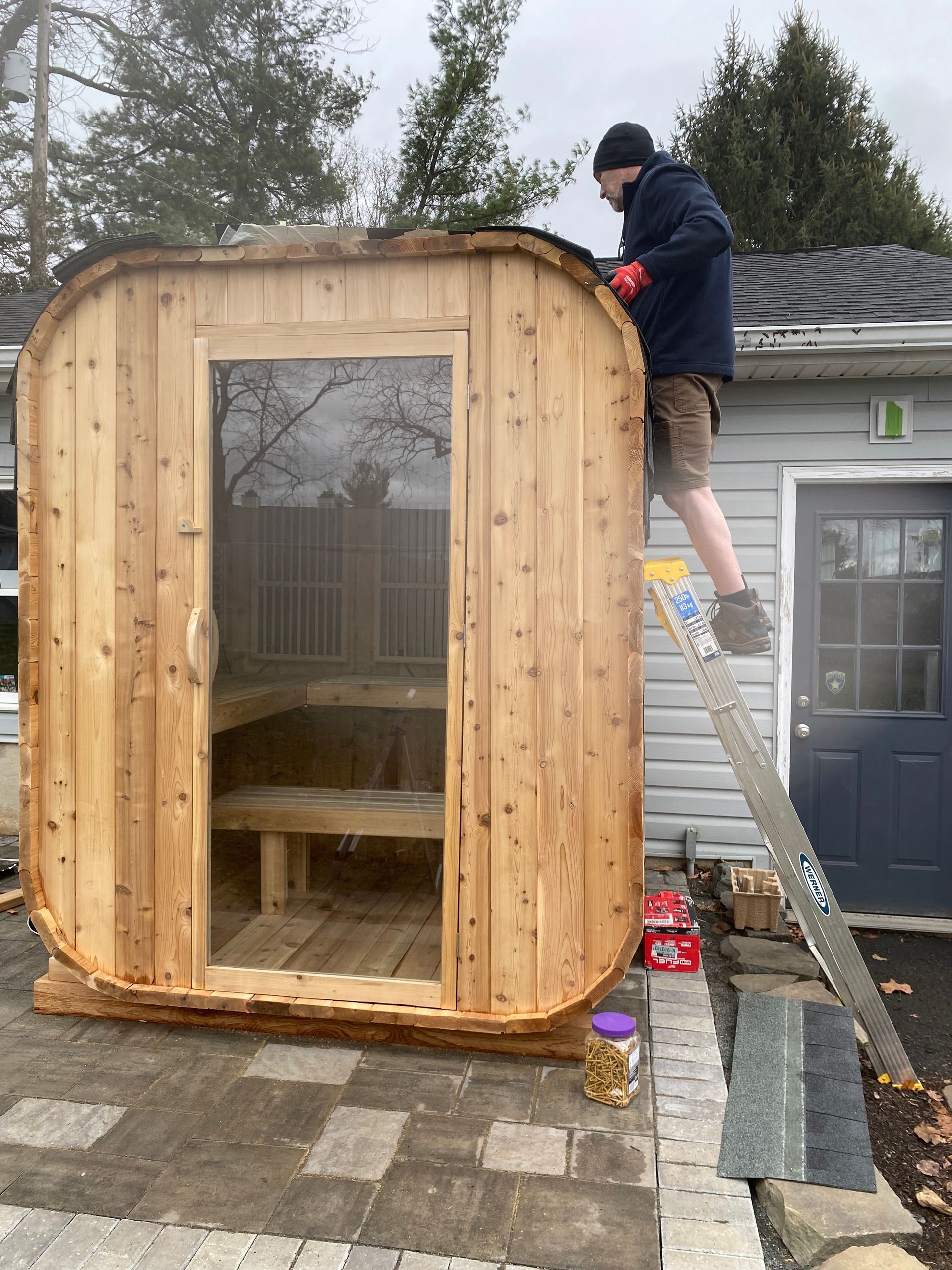 Person standing on a ladder adjusting a small wooden outdoor sauna with a glass door