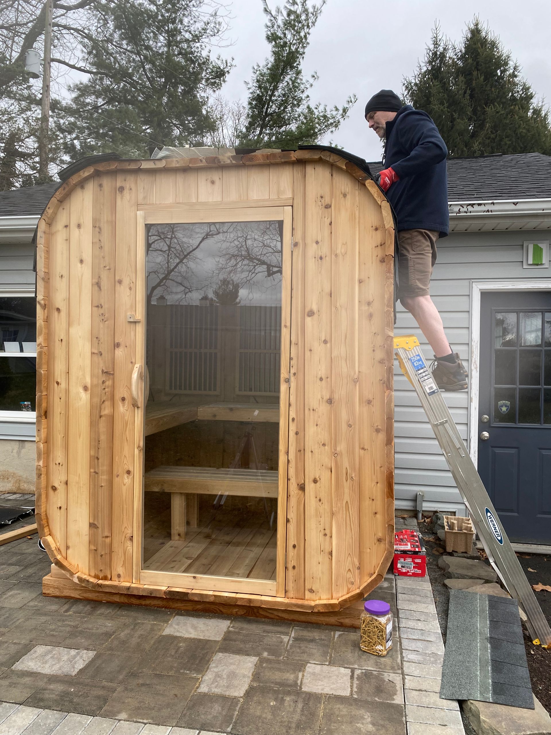 Person on a ladder working on the roof of a small wooden garden shed with a glass door.