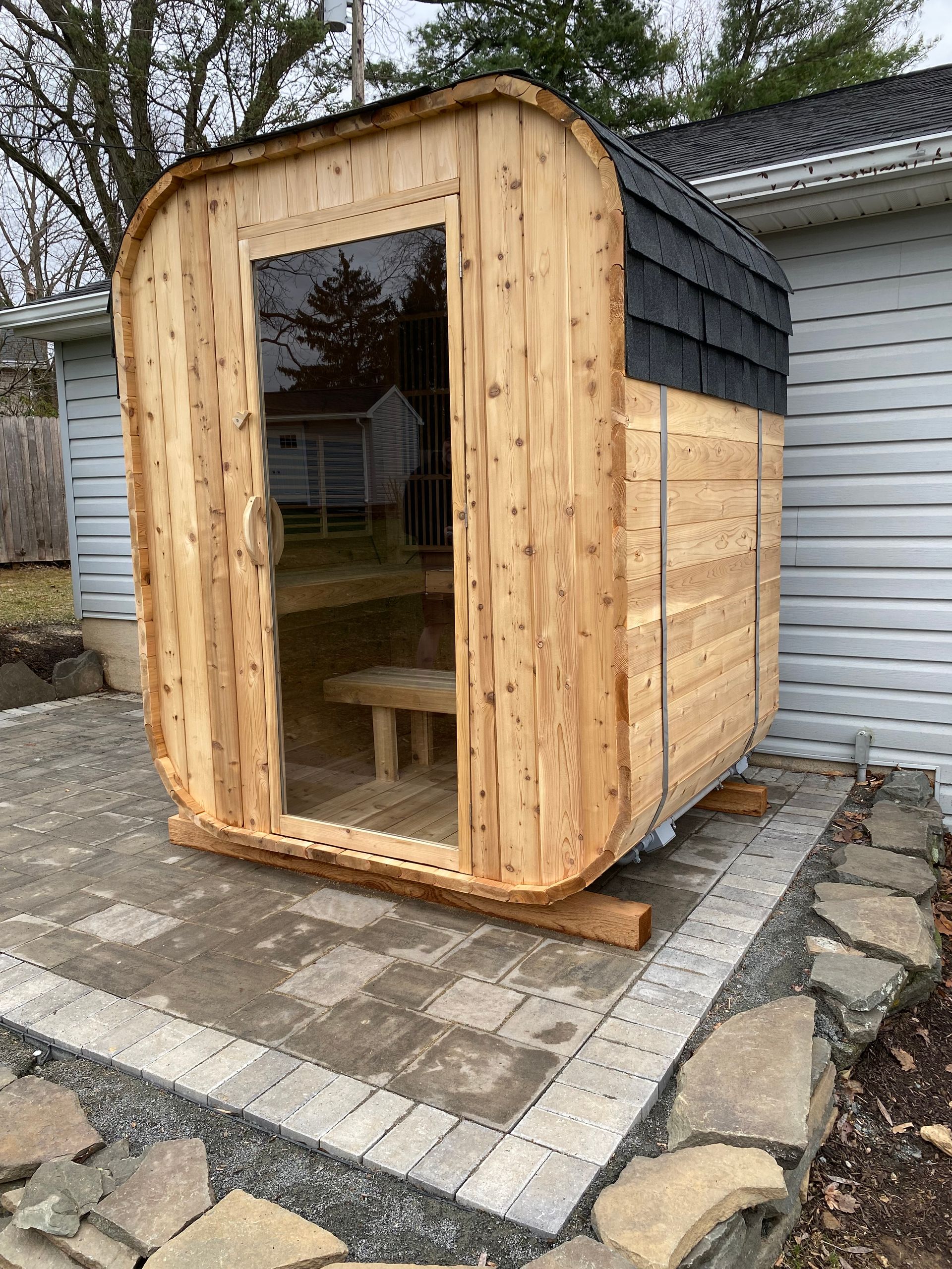 Small wooden sauna cabin with a glass front door, sitting on a paved patio beside a house.