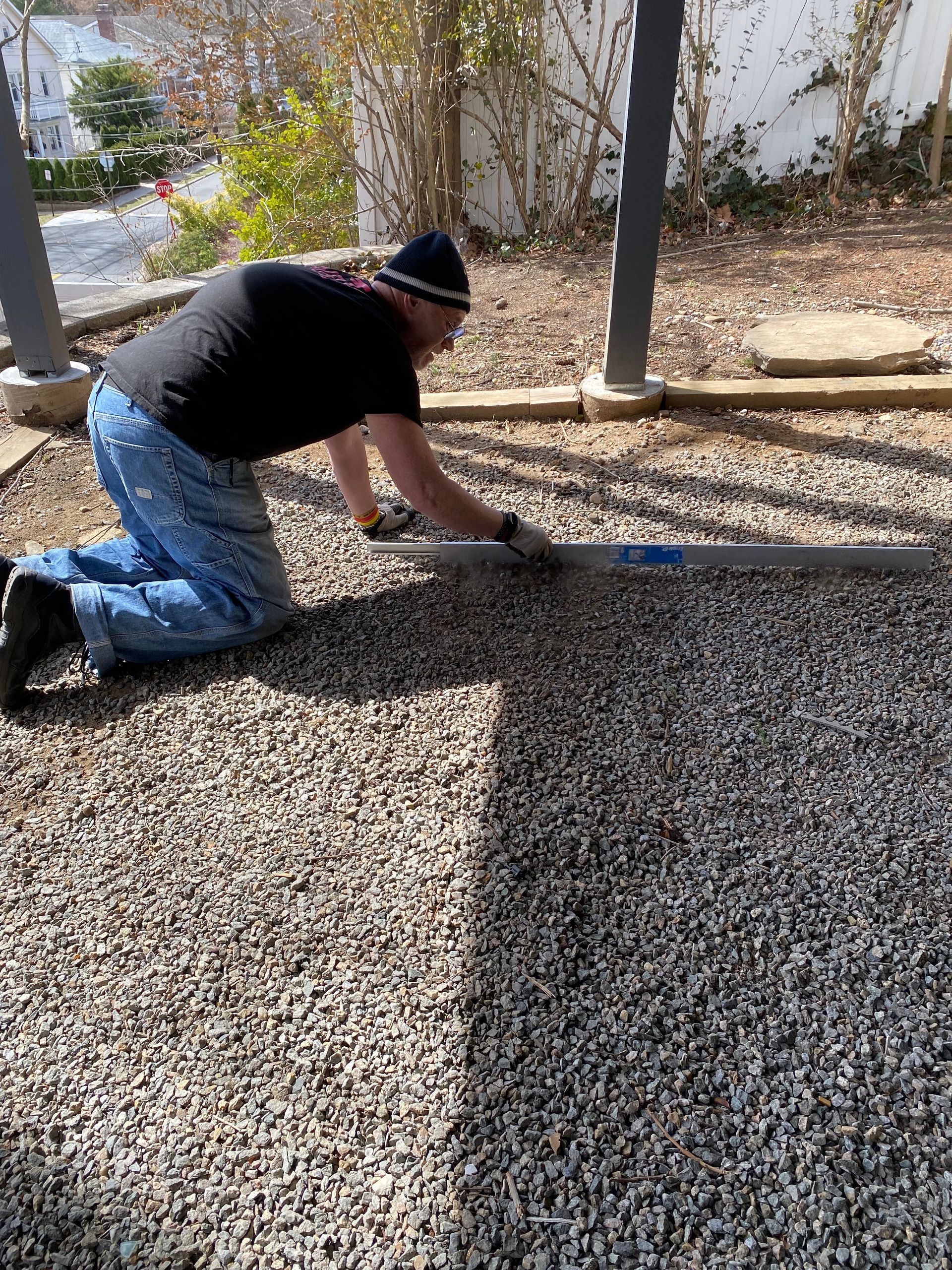 Person kneeling on gravel, smoothing or leveling ground near a patio with a long tool