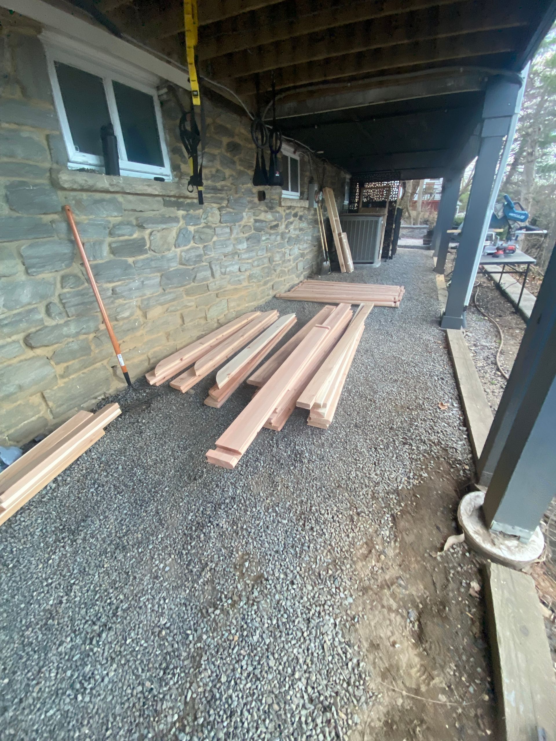 Stacks of lumber on gravel beside a stone wall under a covered walkway.