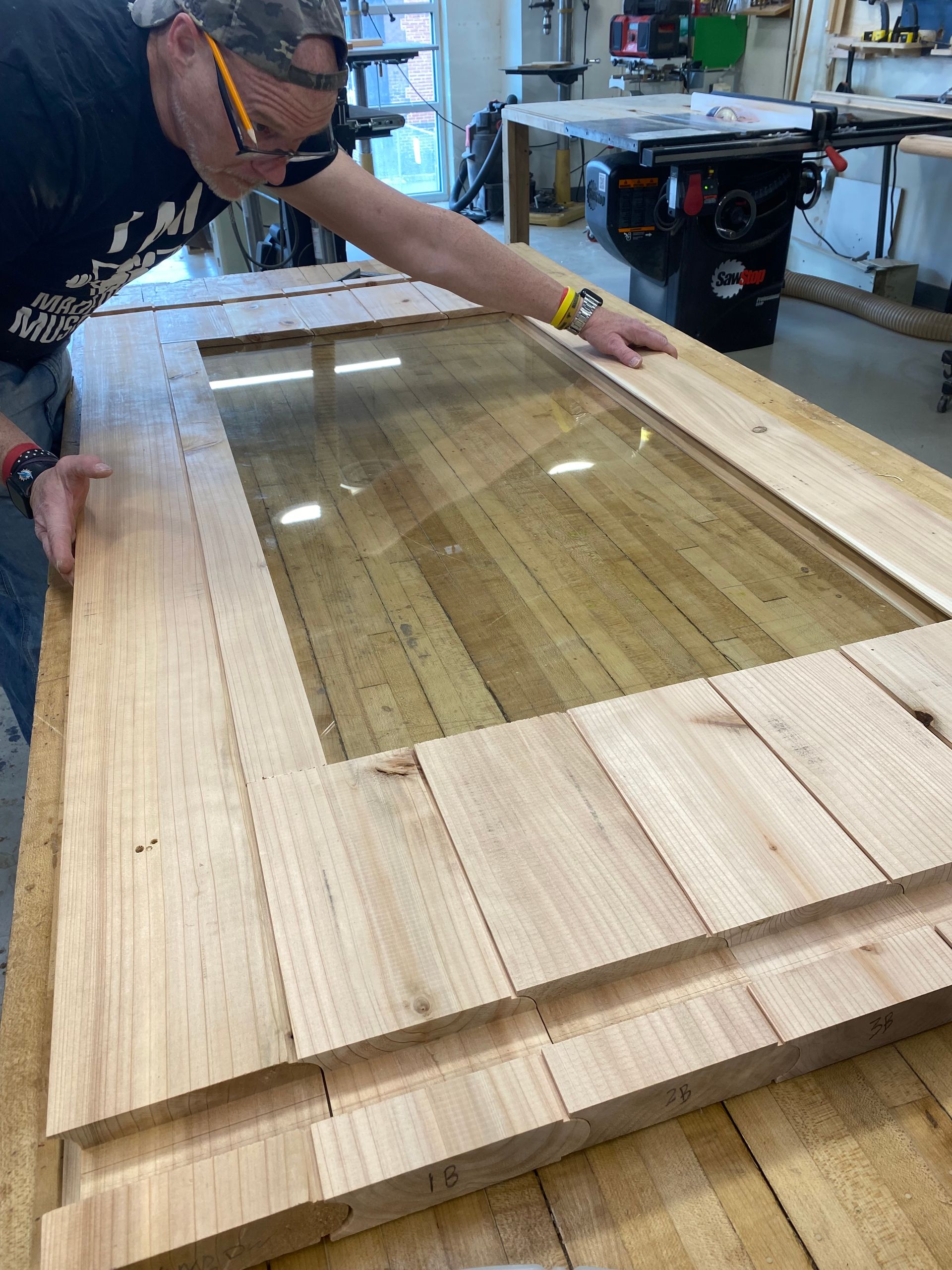 Worker leaning over a wooden frame on a workshop table, fitting a glass panel into place.
