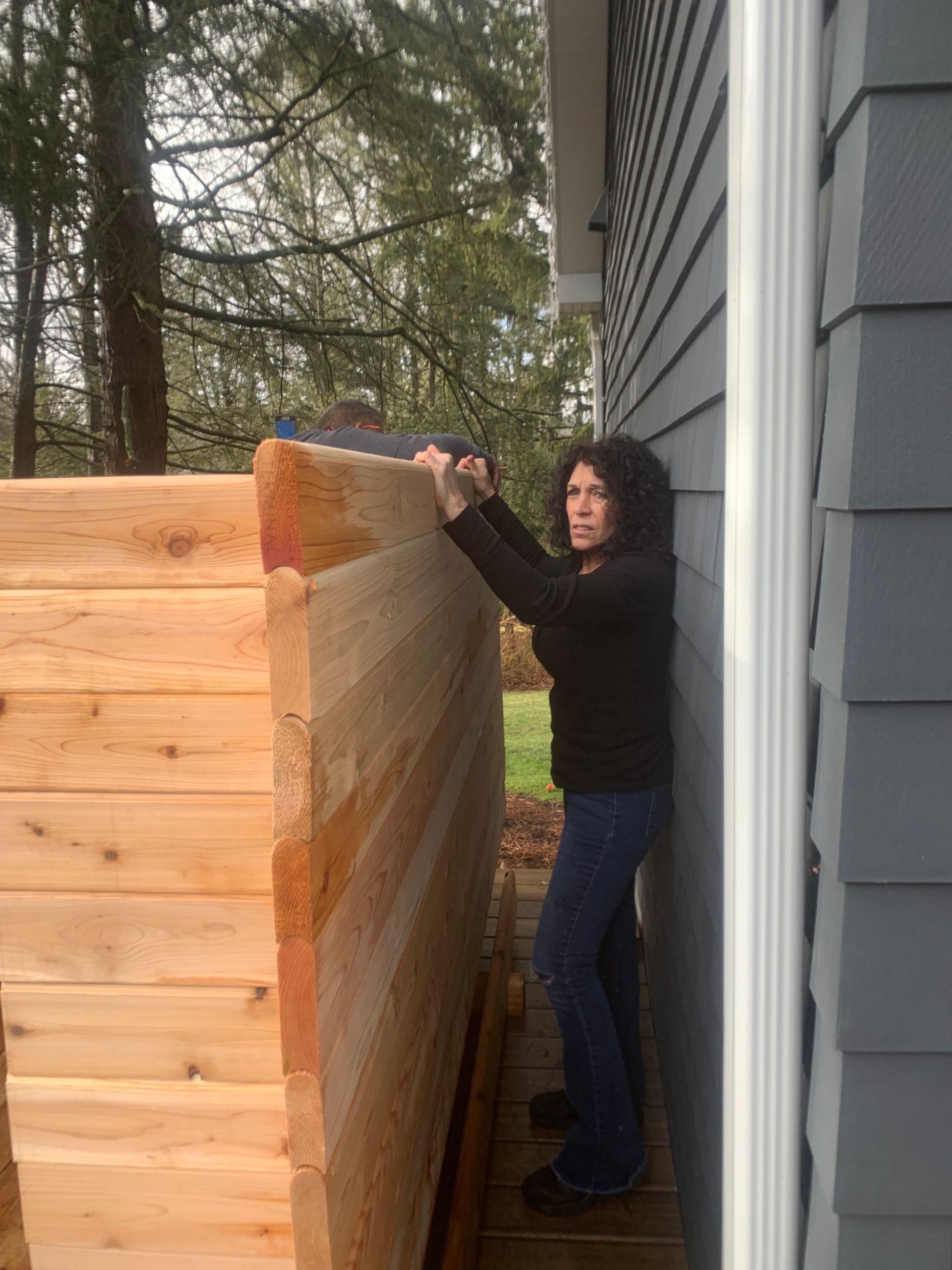 Person in black jacket moving a large wrapped bundle beside a house and wooden structure outdoors.