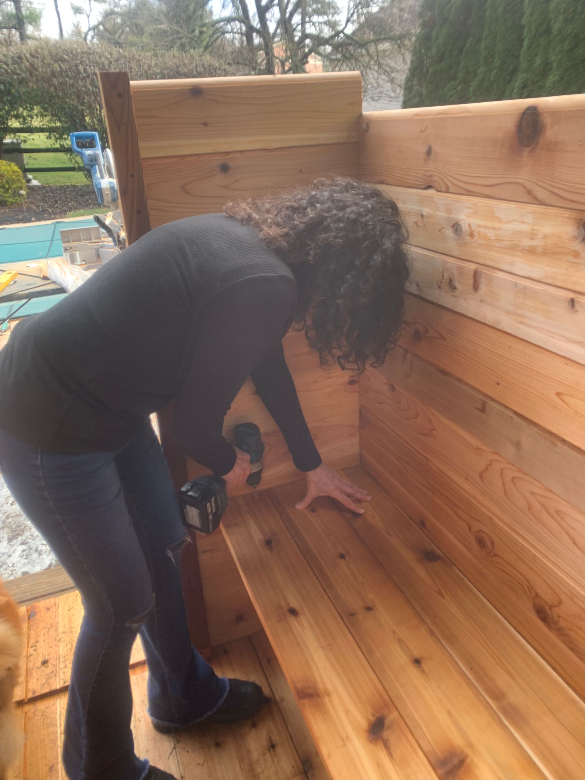 Person bending over using a drill on a wooden bench in an outdoor setting