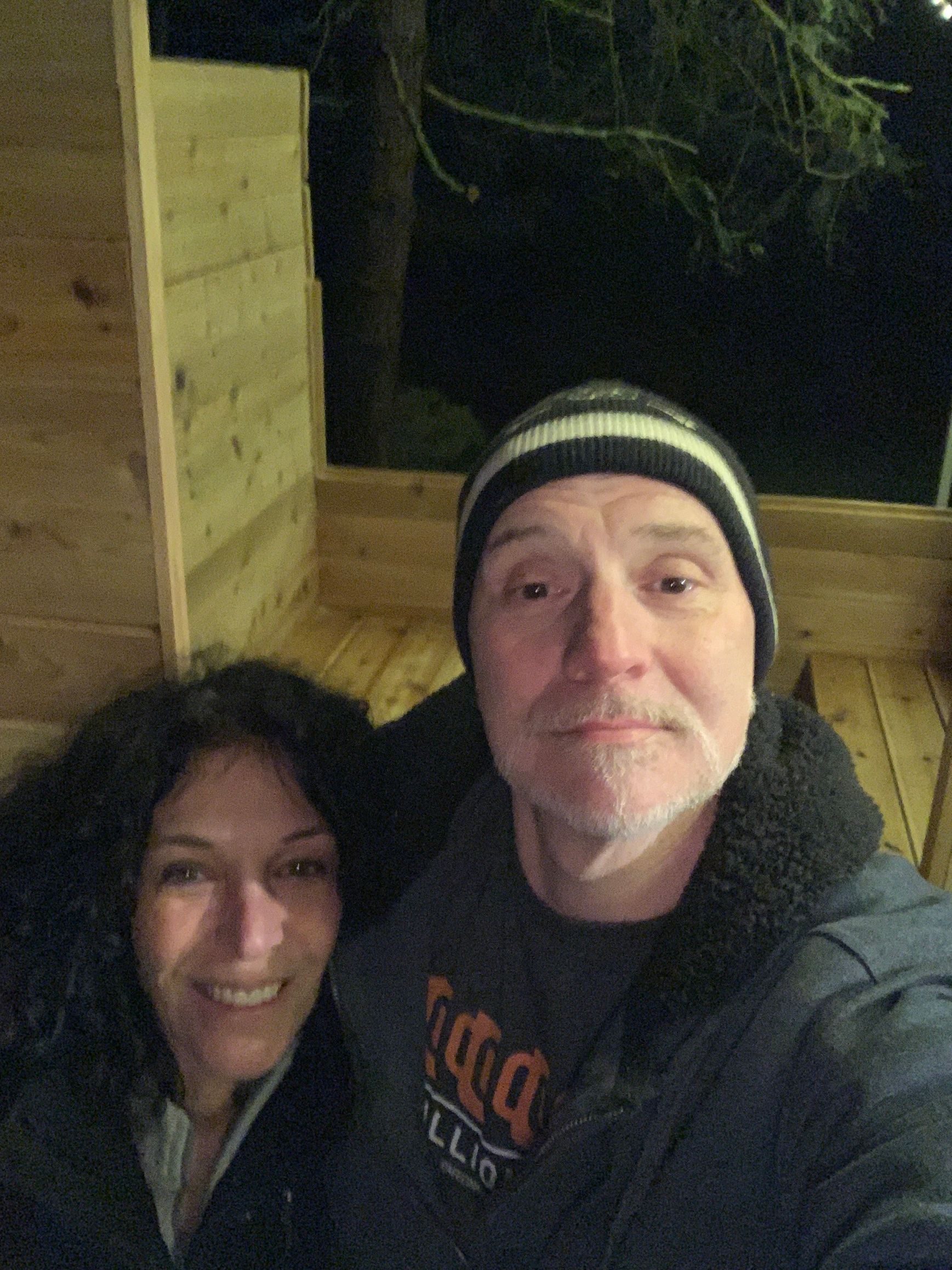 Two people smiling in a dimly lit outdoor selfie at night, standing beside a wooden wall.