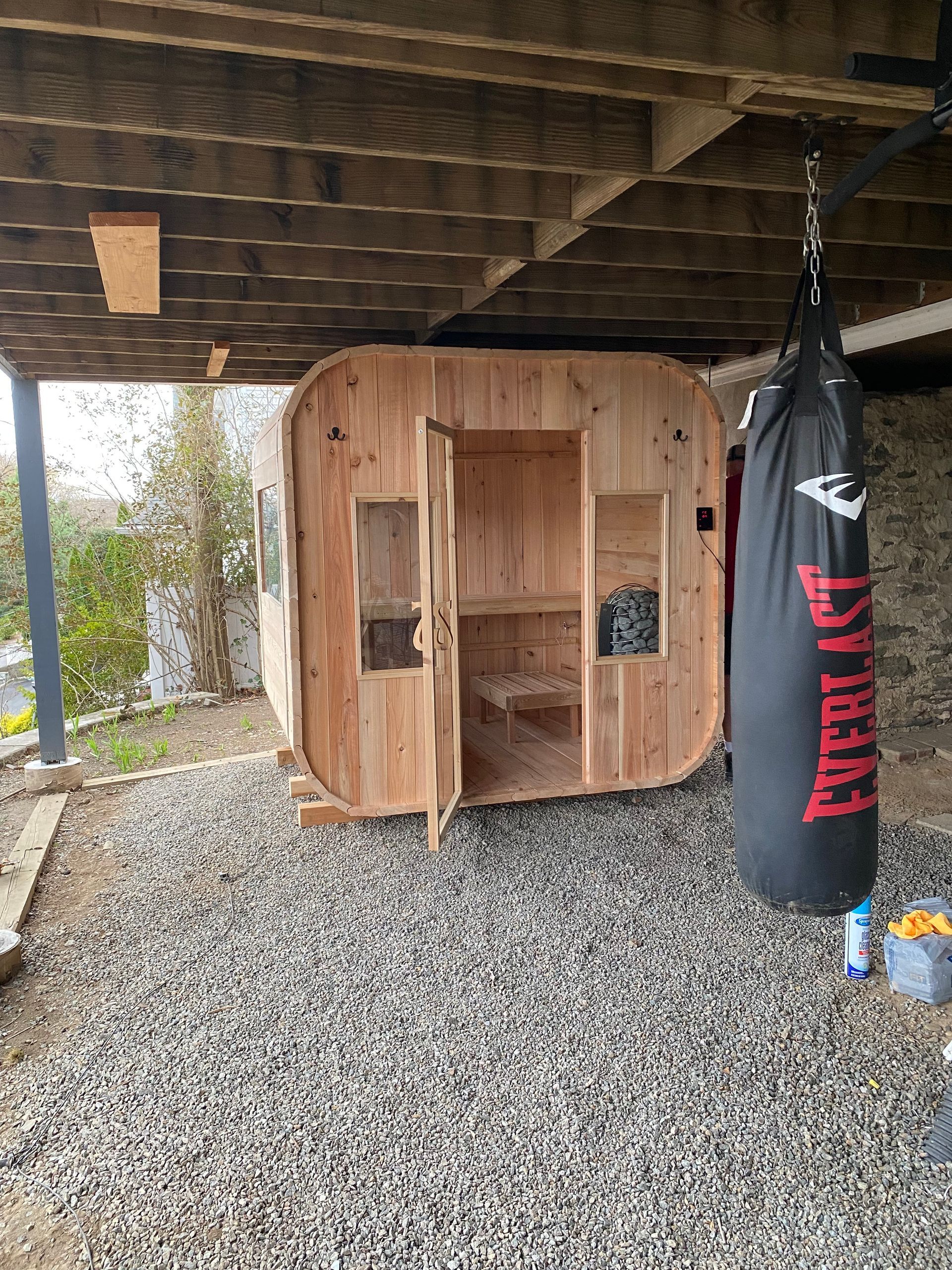 Wooden sauna shed under a covered patio, with a black punching bag hanging beside it.
