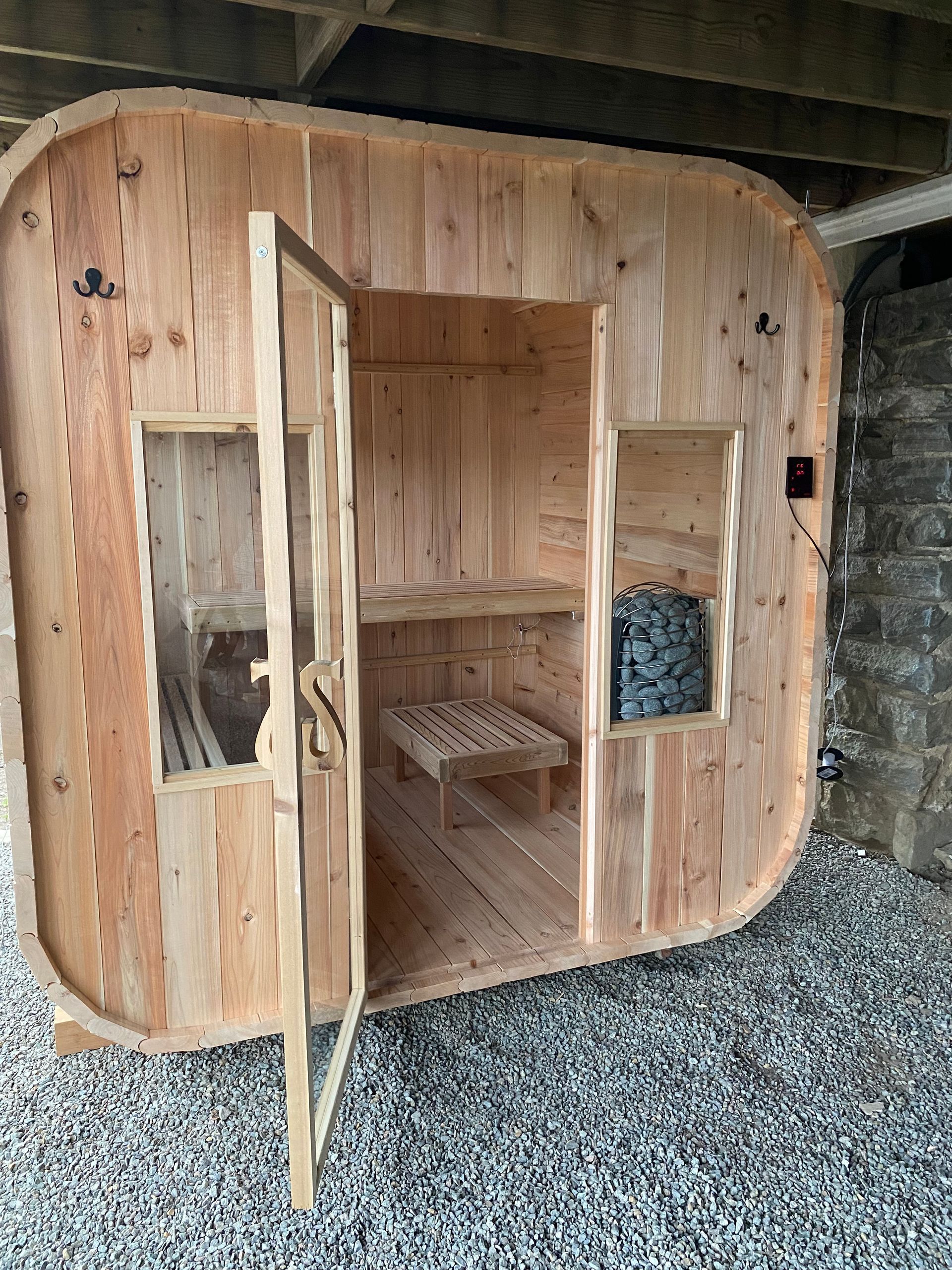 Small round wooden sauna cabin with an open door, showing a bench inside on gravel ground.