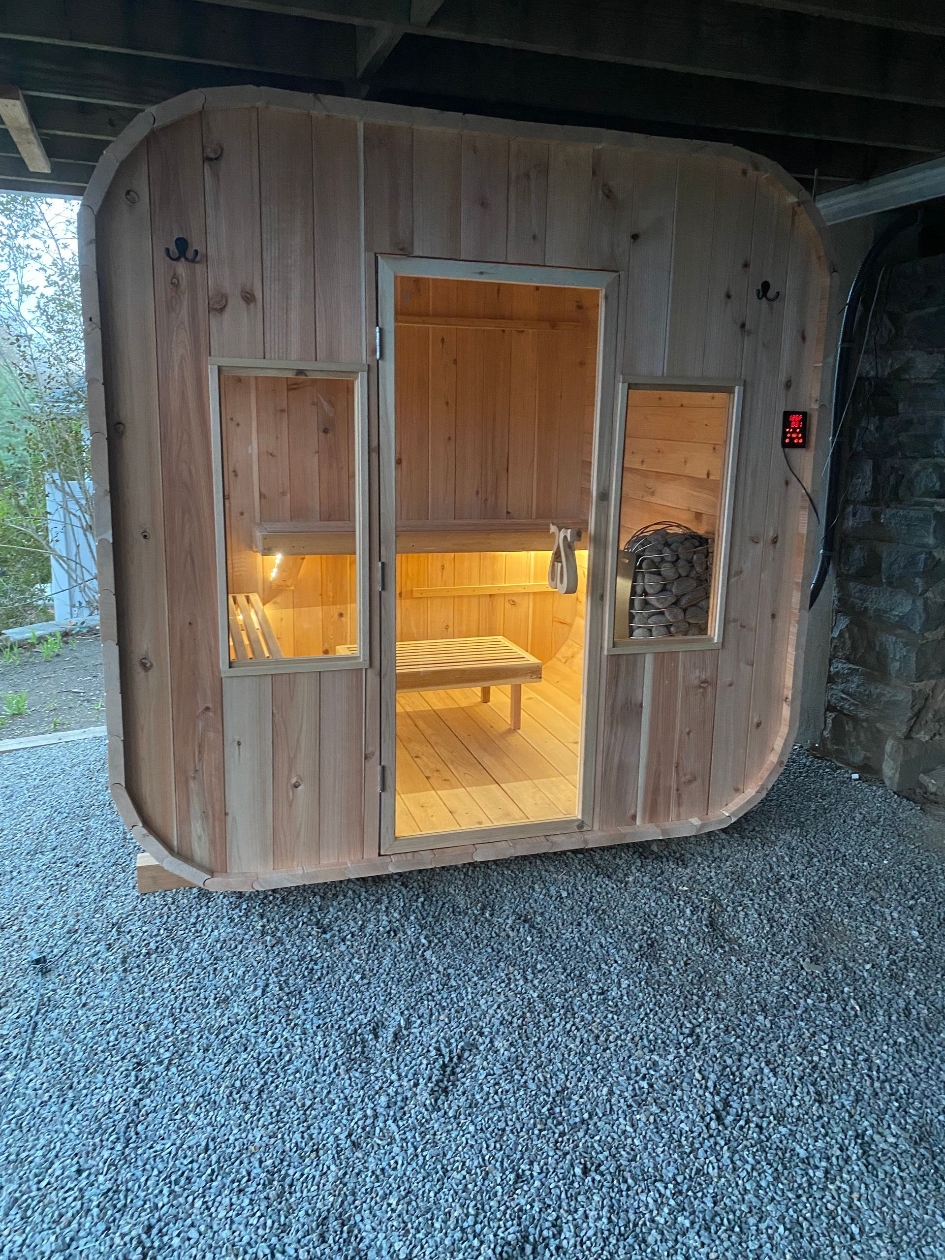 Small wooden sauna cabin with open door, warm interior lights, and gravel ground outside