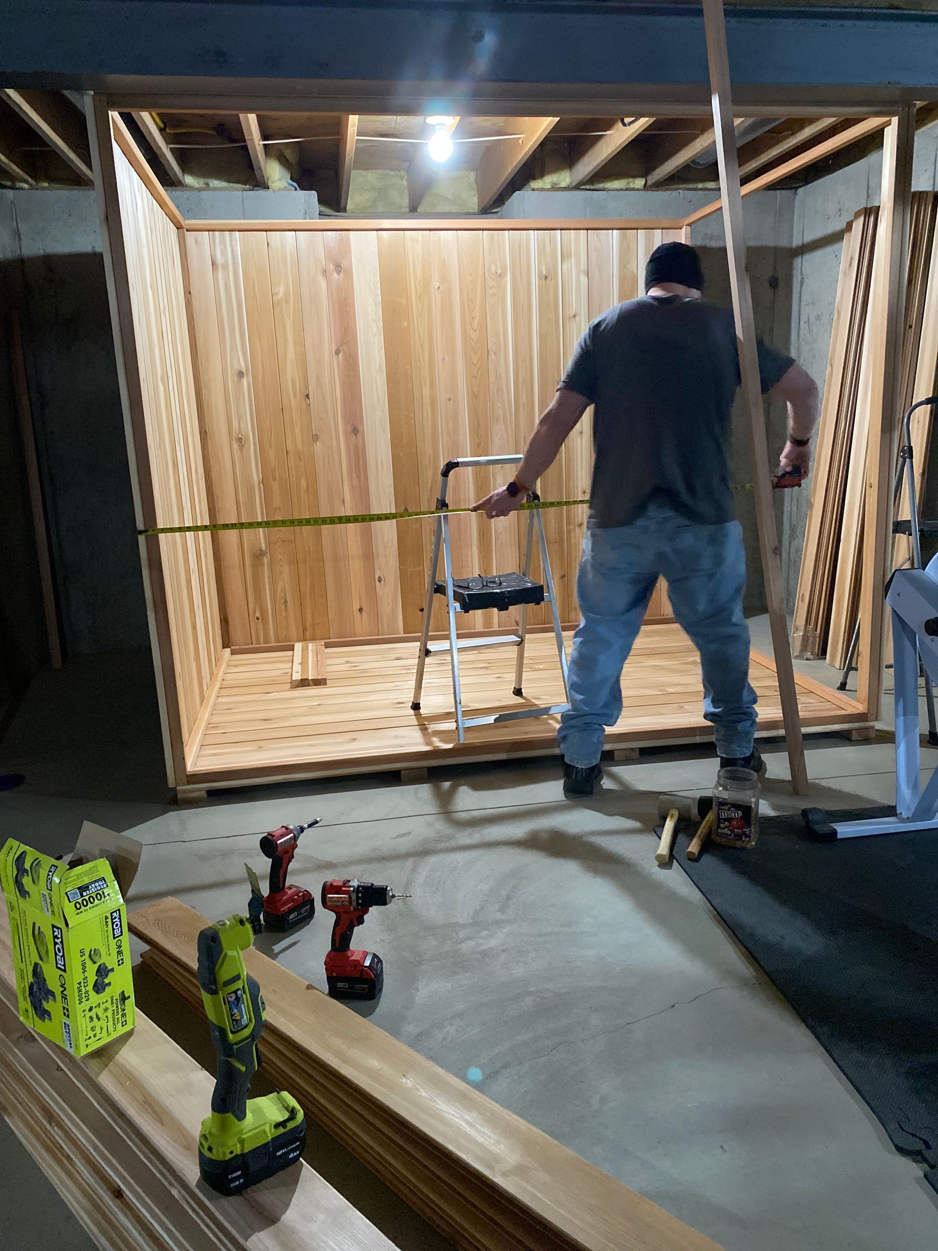 Person assembling a wood-framed structure in a workshop, with tools and lumber on the floor.