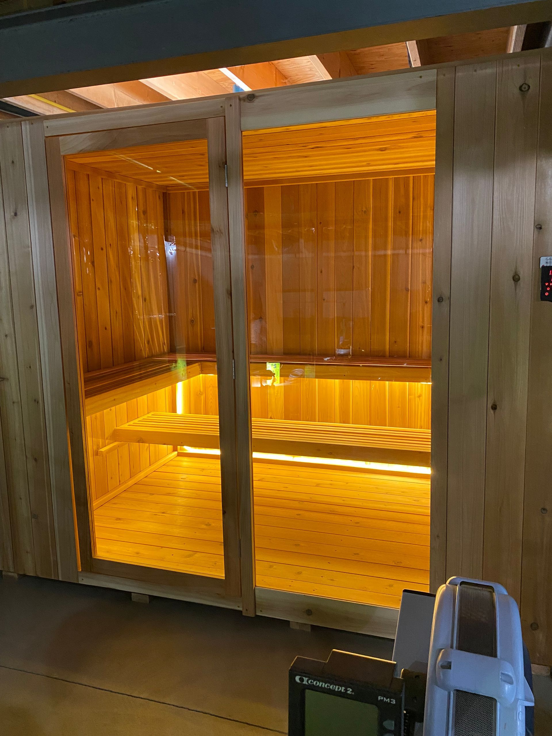 Lit wooden sauna room with glass doors and benches, viewed from an airport-like corridor.