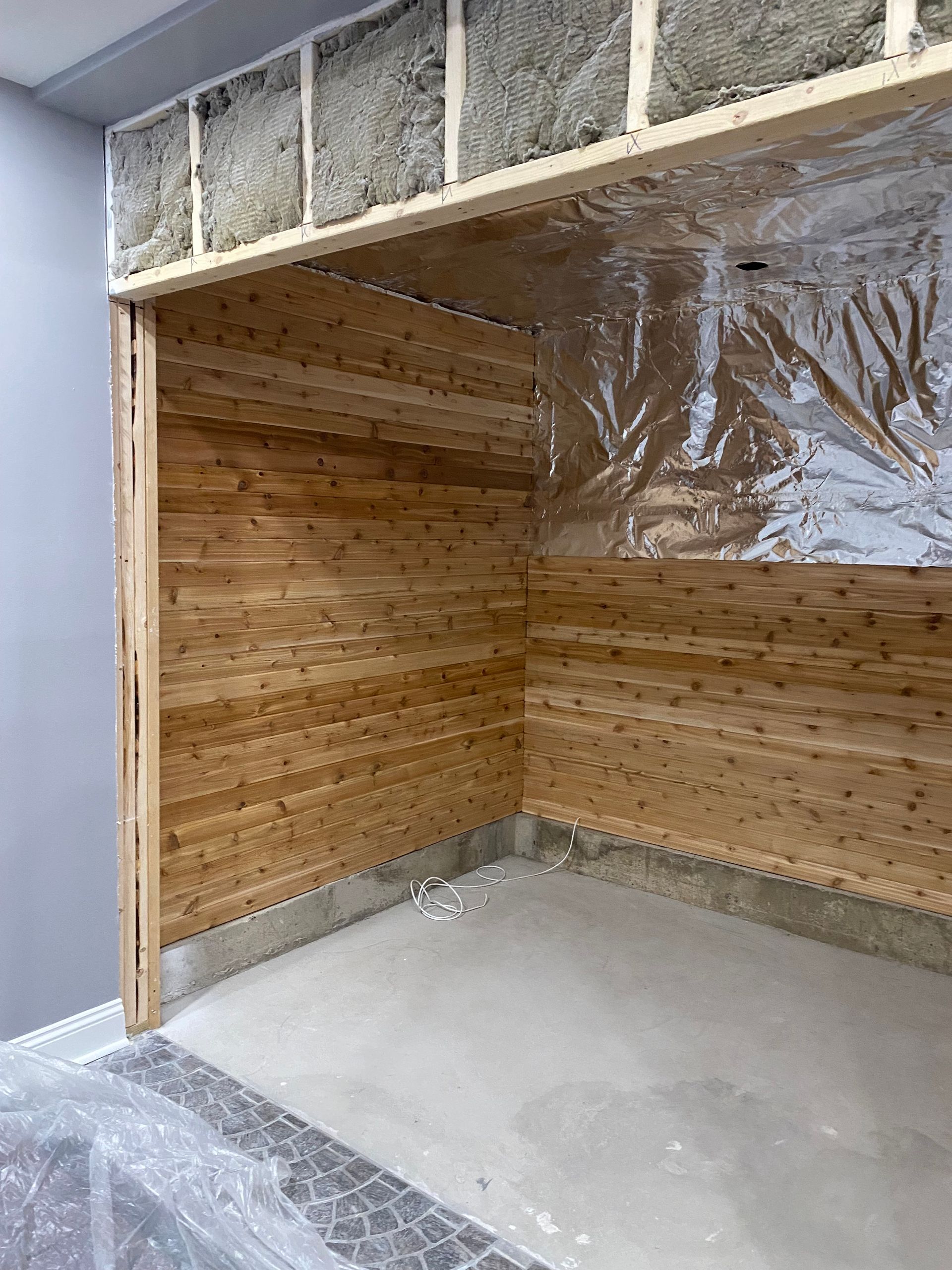 Empty corner of a room with wood-paneled walls and plastic sheeting overhead, under construction.