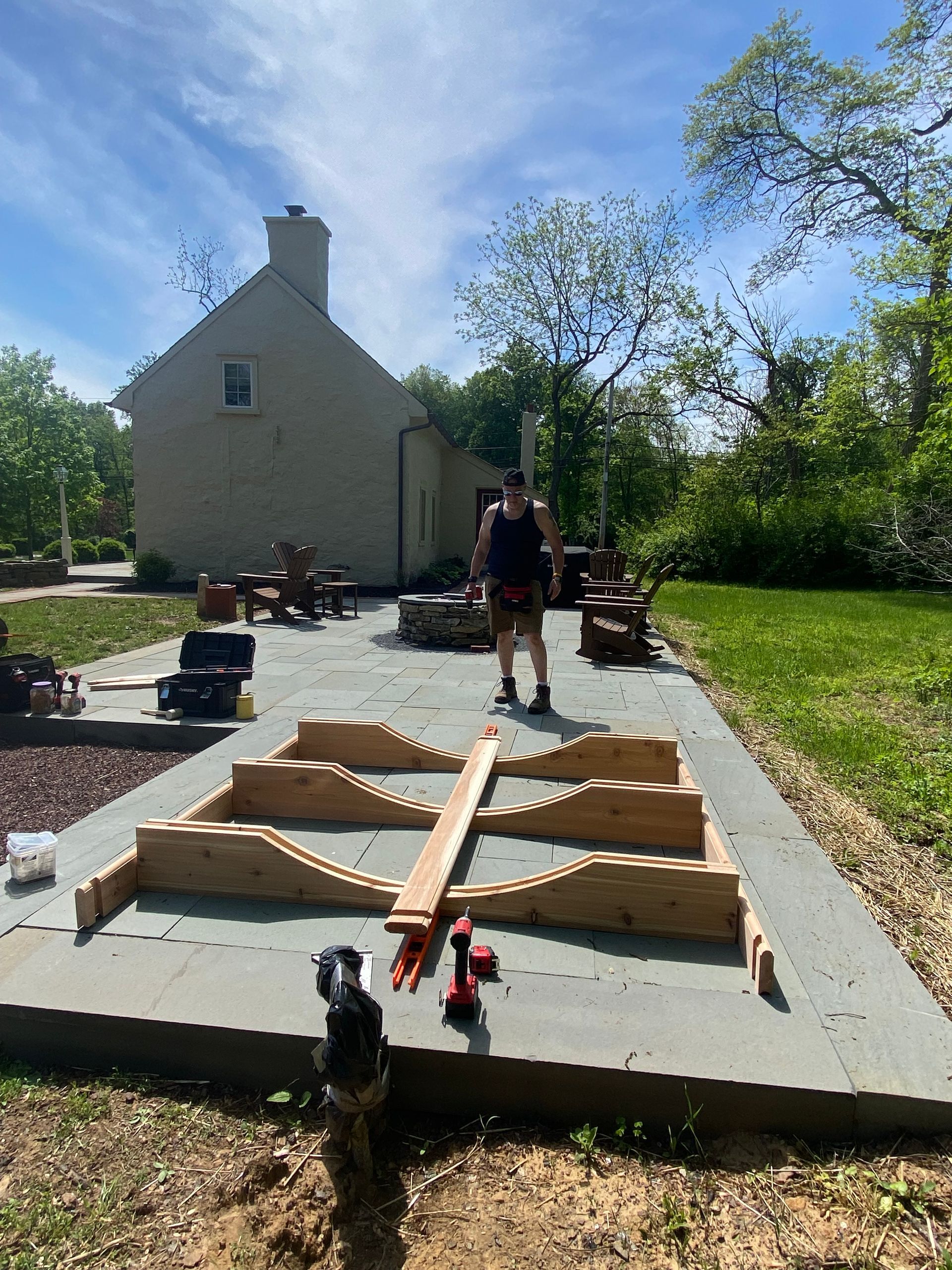 Man working on a wooden frame on a concrete patio beside a house, with tools and gravel in the yard.