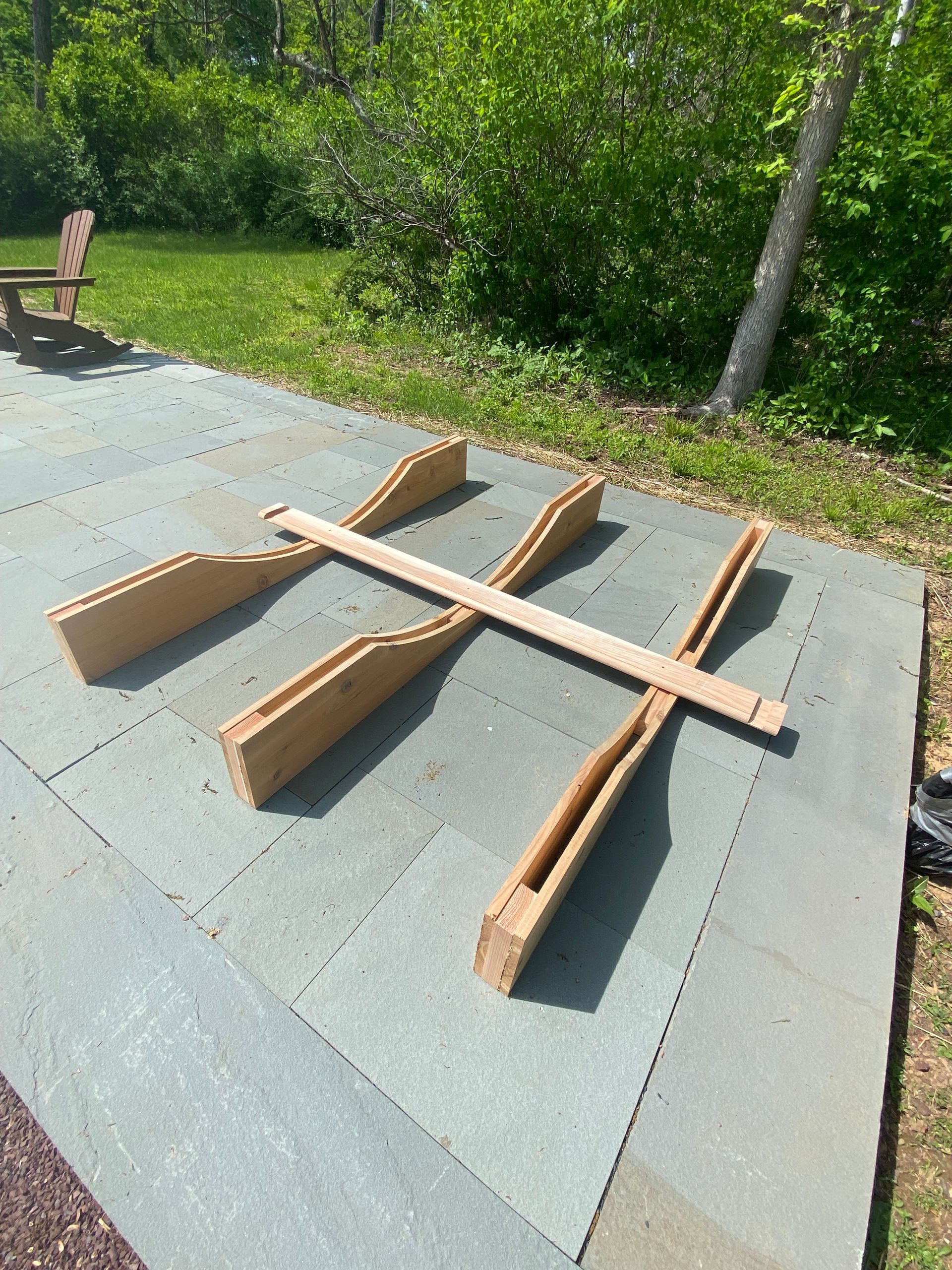 Several wooden beams laid out on a concrete slab beside a grassy area and trees