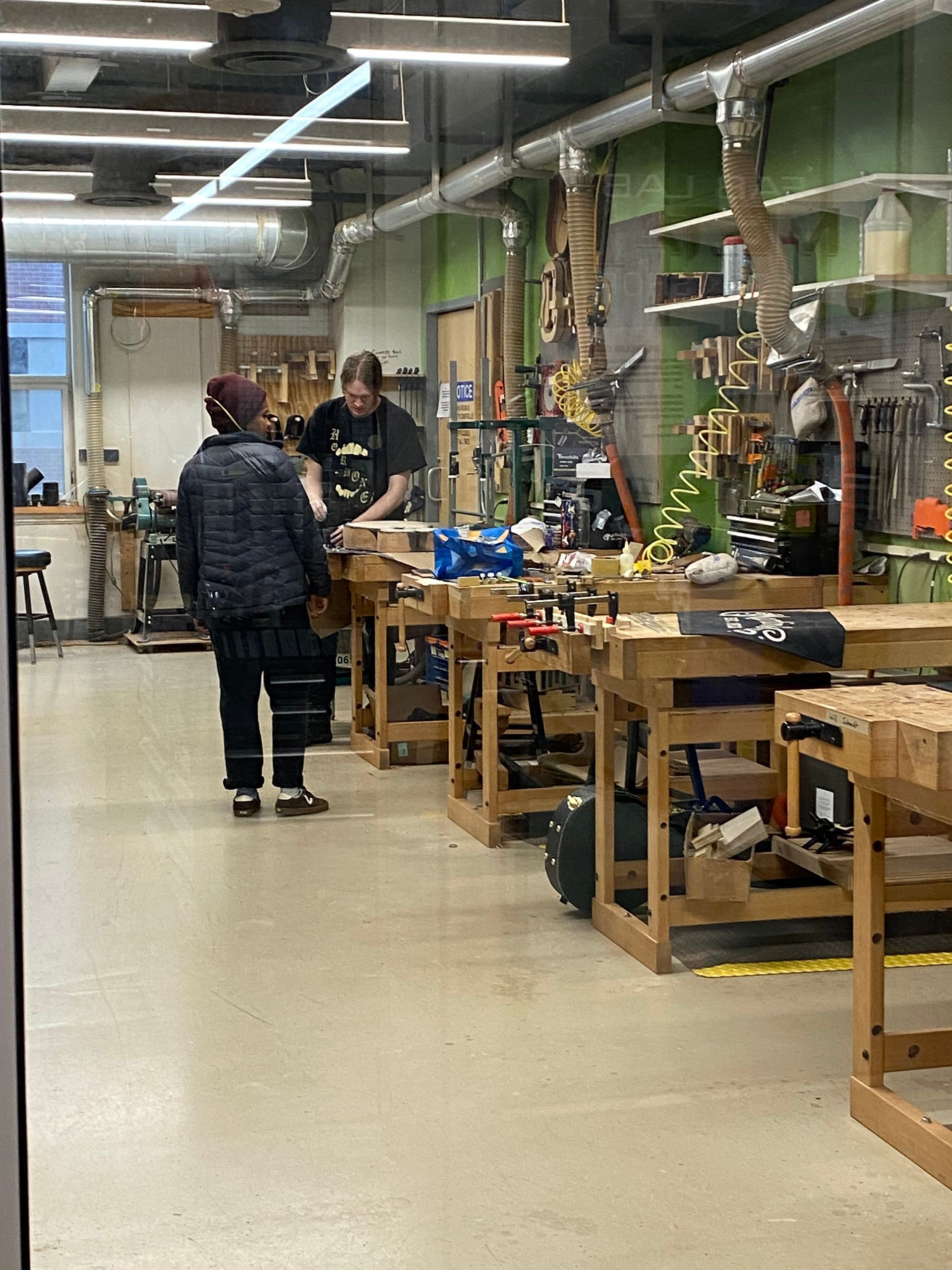 Workshop aisle with two people near wooden workbenches and tools under bright overhead lights
