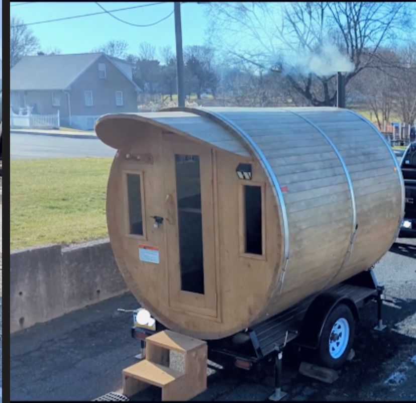 Small wooden barrel-shaped tiny house on a trailer in a driveway, with smoke from a chimney.